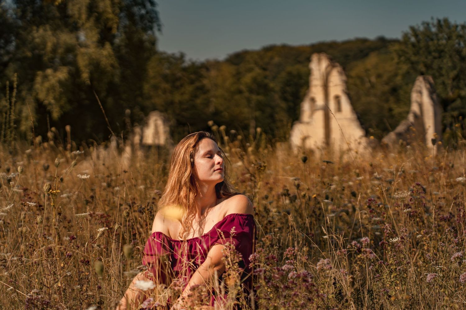 Woman in red dress sitting in golden meadow with ancient ruins and trees in background on sunny day.