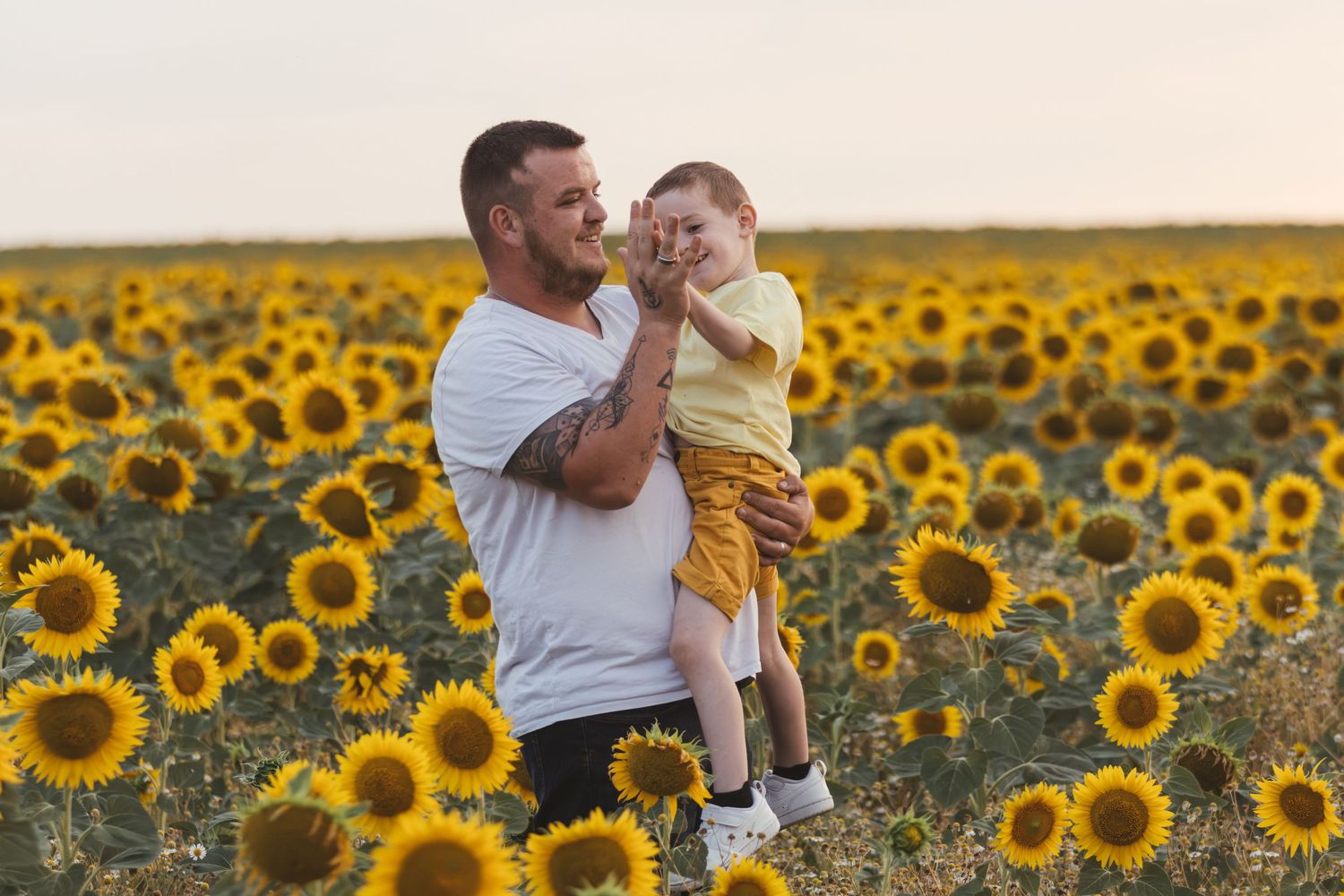 Father holding young son in a vibrant sunflower field at sunset, sharing a joyful moment together.
