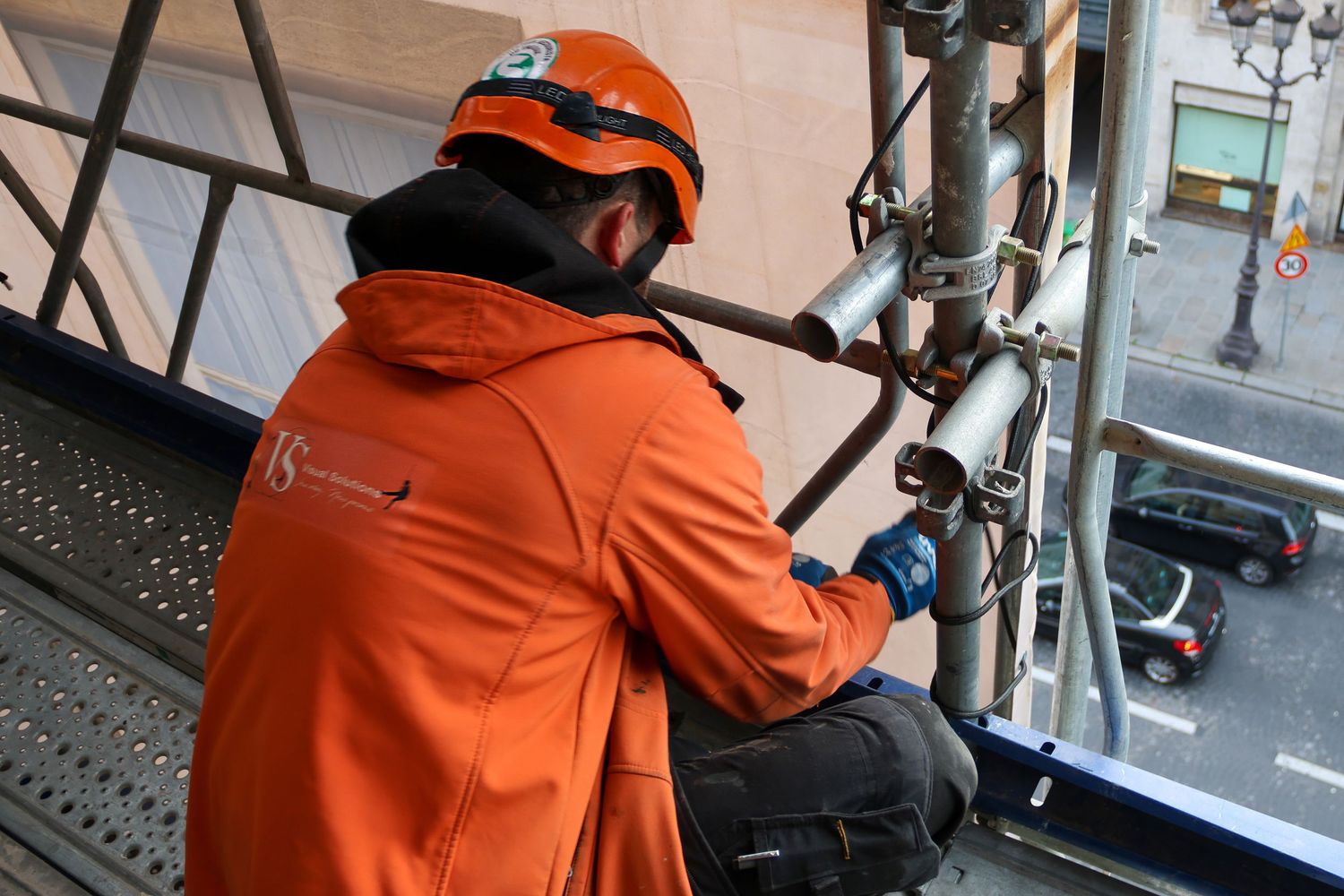 Worker in orange jacket and helmet performing maintenance on scaffolding structure at height.