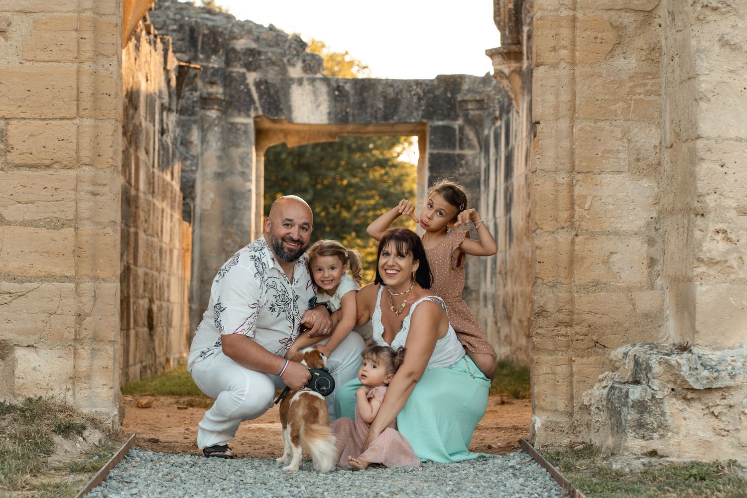 A happy family of five poses with their dog among stone ruins at sunset, parents holding young children.