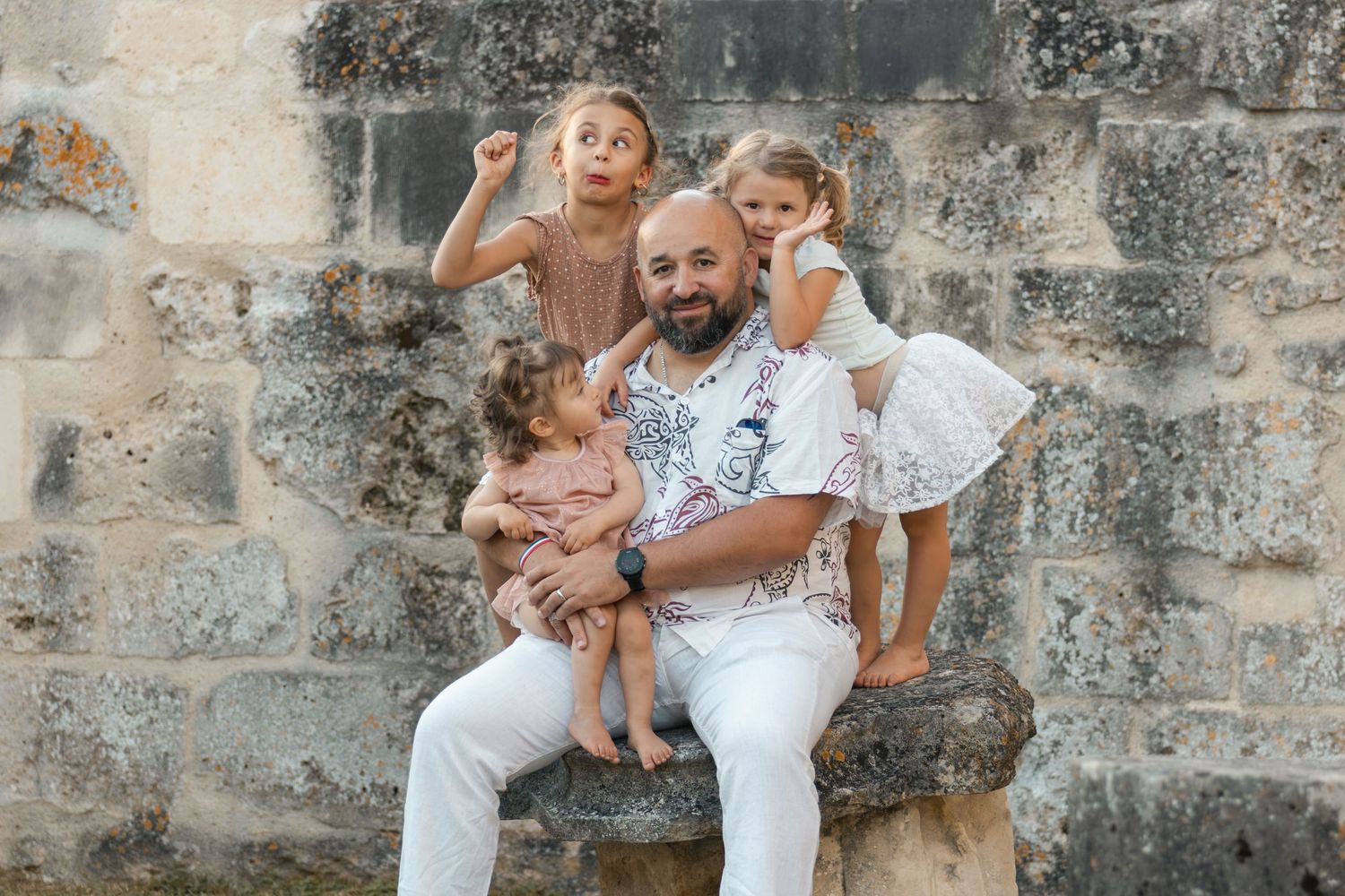 A smiling man in white sits on a stone bench holding a toddler, with two young girls posing near an old stone wall.