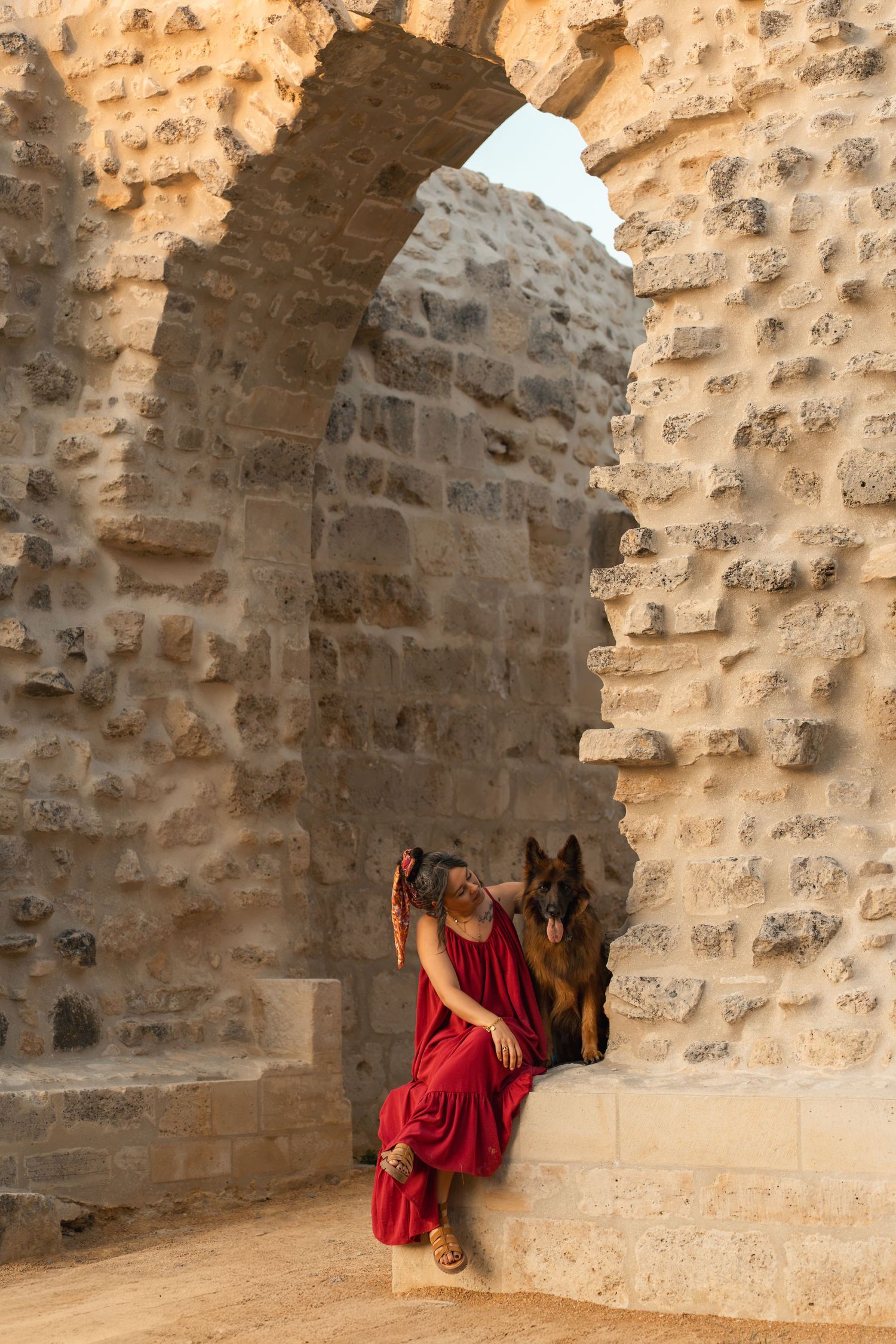 Woman in red dress sitting with German Shepherd dog in ancient stone archway ruins.