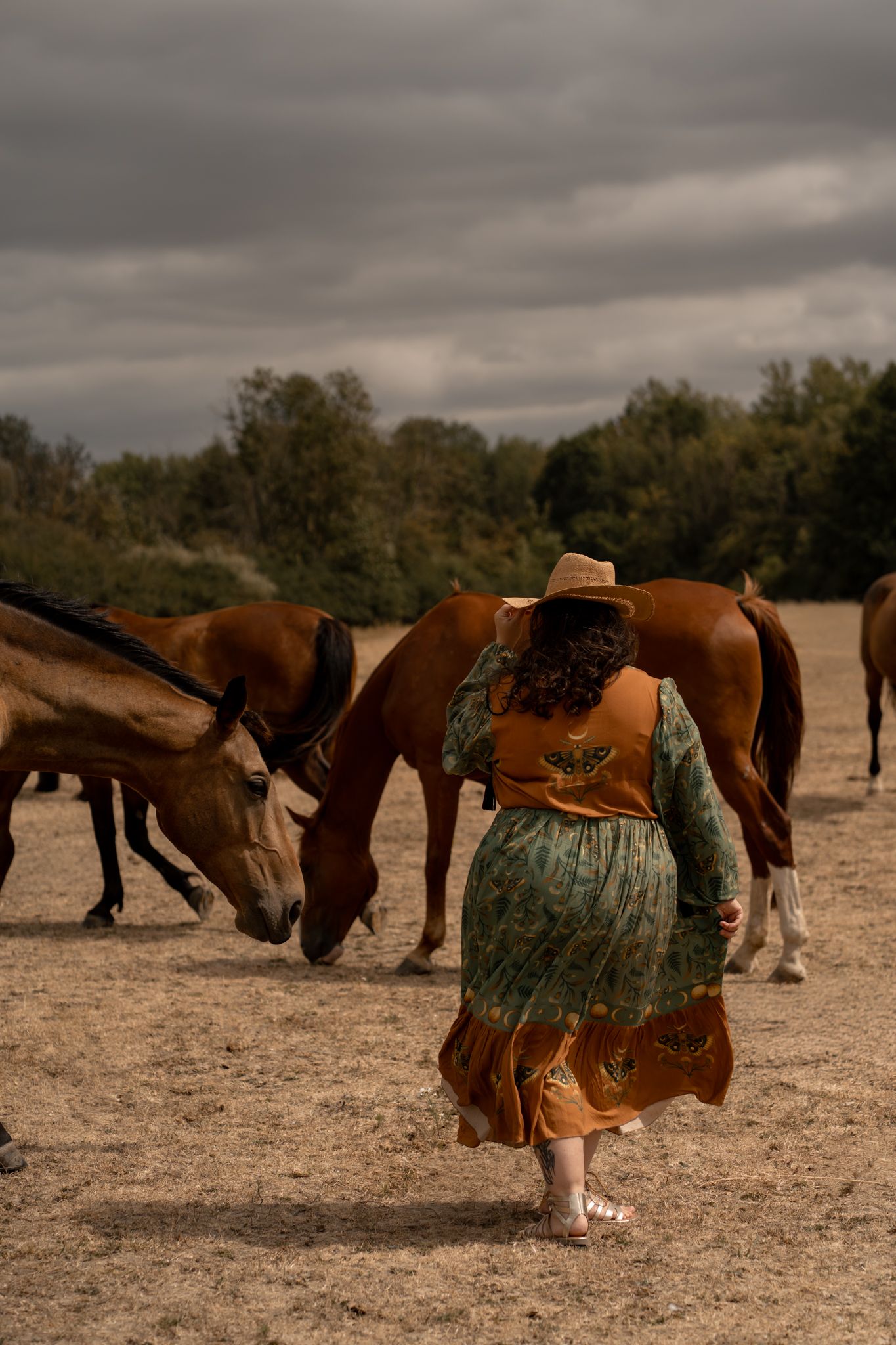 A woman in a green floral dress and hat walks barefoot among grazing brown horses on a dusty field.