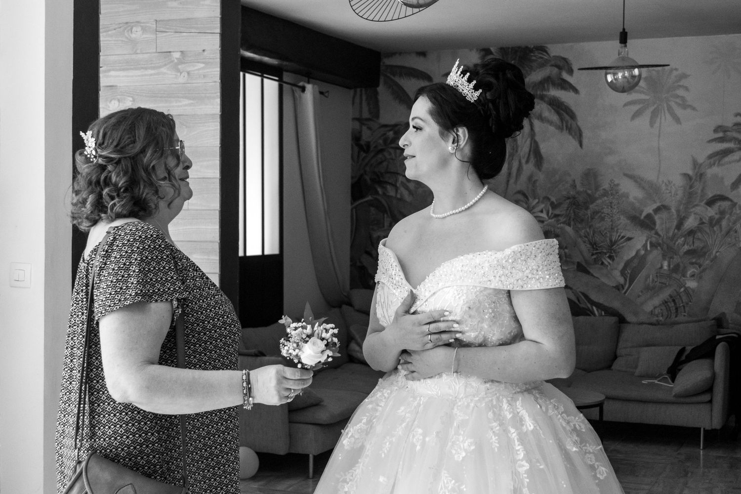 Black and white photo of a bride in a ball gown and tiara speaking with a woman holding a small bouquet indoors.