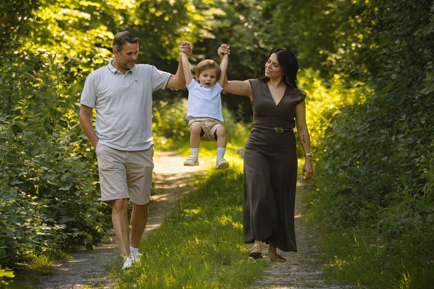 Family with parents swinging young toddler child by hands on sunny green tree-lined garden path.