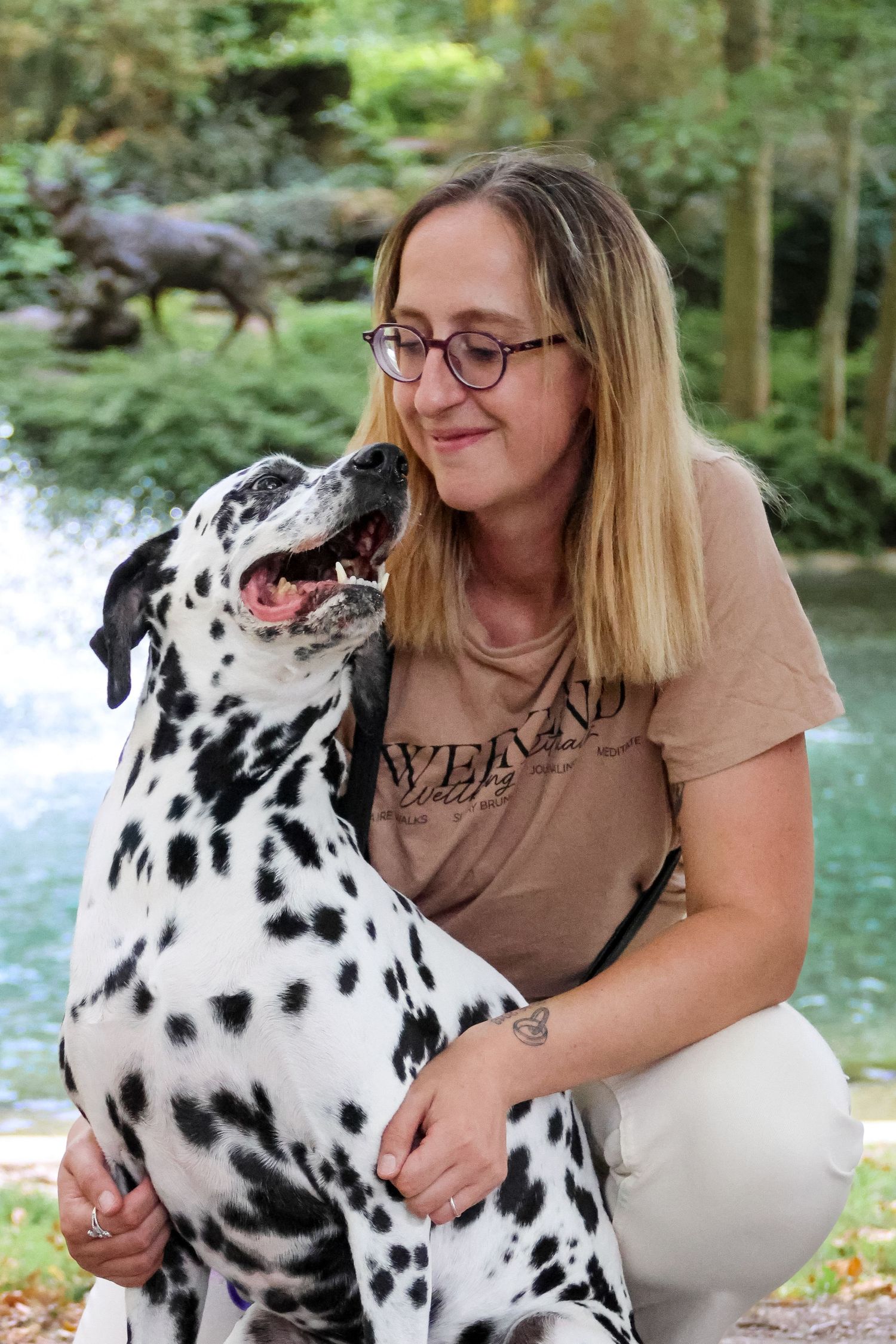 Woman with glasses crouching beside a Dalmatian dog near a pond with green trees in the background.