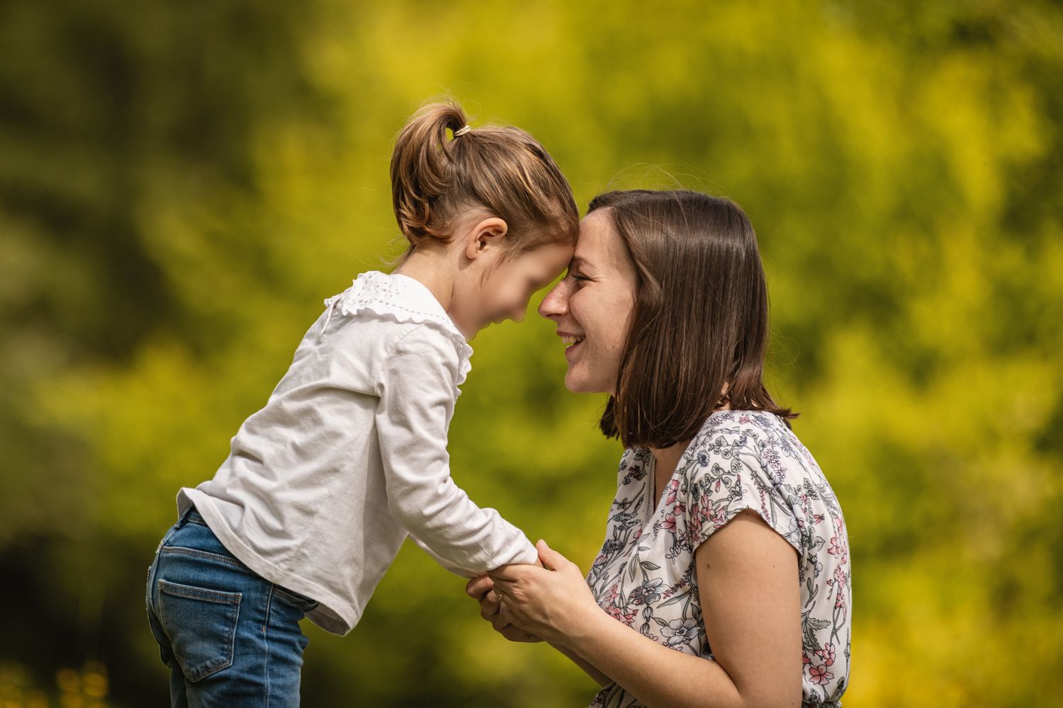 A smiling mother and young daughter touch foreheads joyfully outdoors with lush green trees in the background.