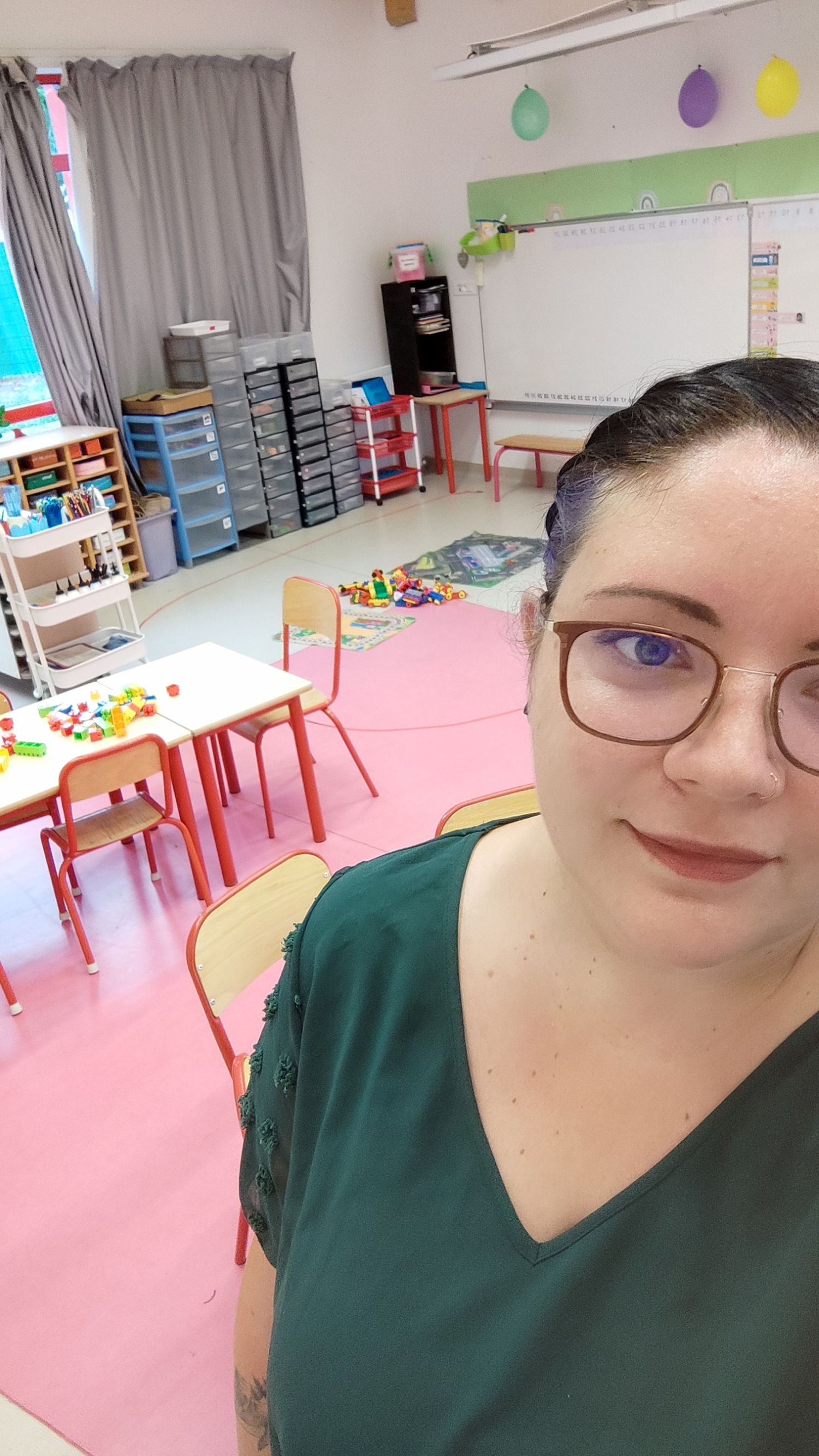 Woman with glasses in green top takes selfie in colorful classroom with small chairs, tables, and toys.