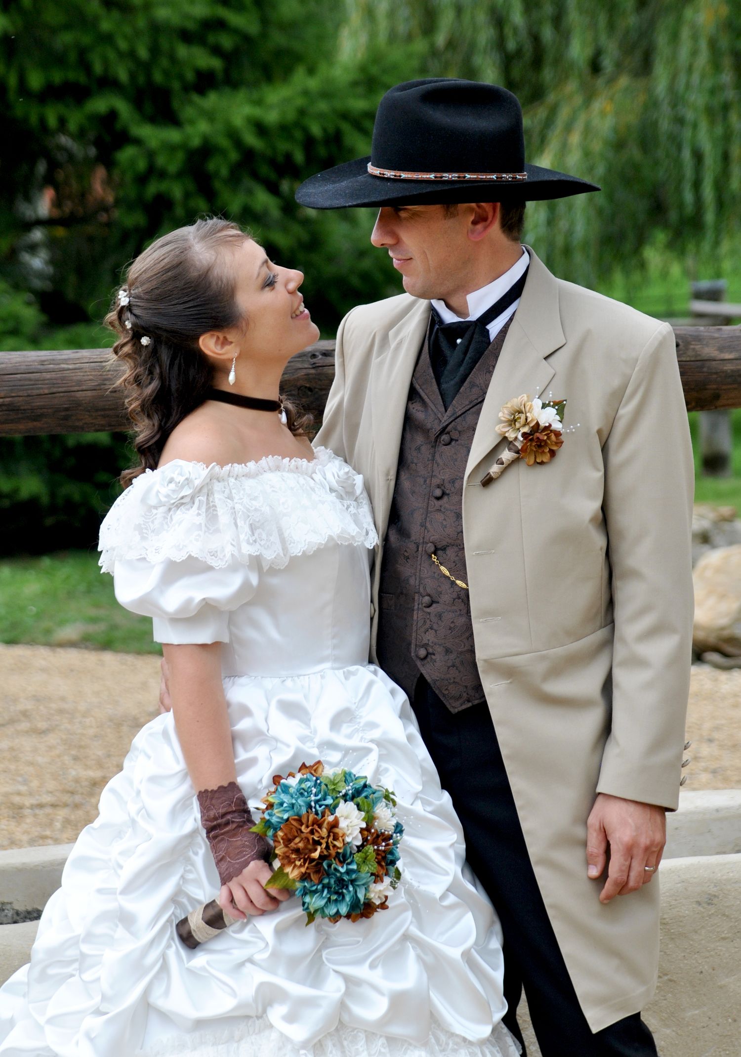 Western bride and groom gazing at each other, she holds teal bouquet, he wears black cowboy hat and tan suit.
