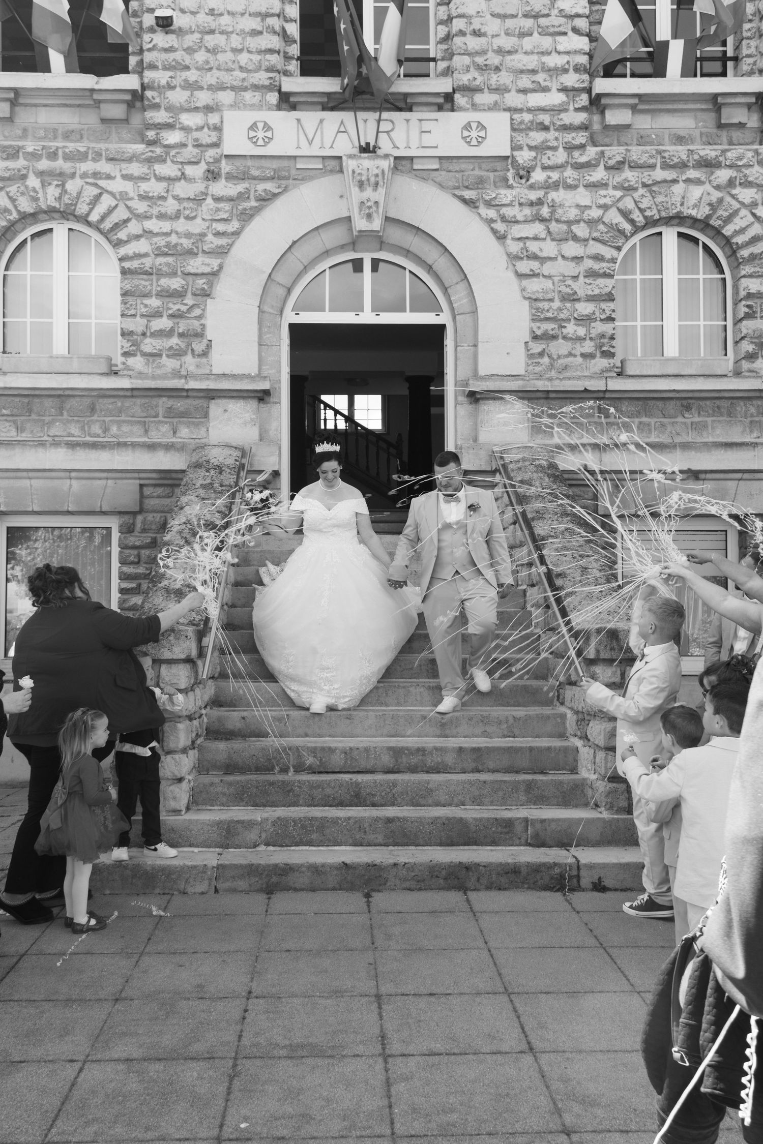 Black and white photo of bride and groom descending mairie steps, greeted by guests throwing streamers.