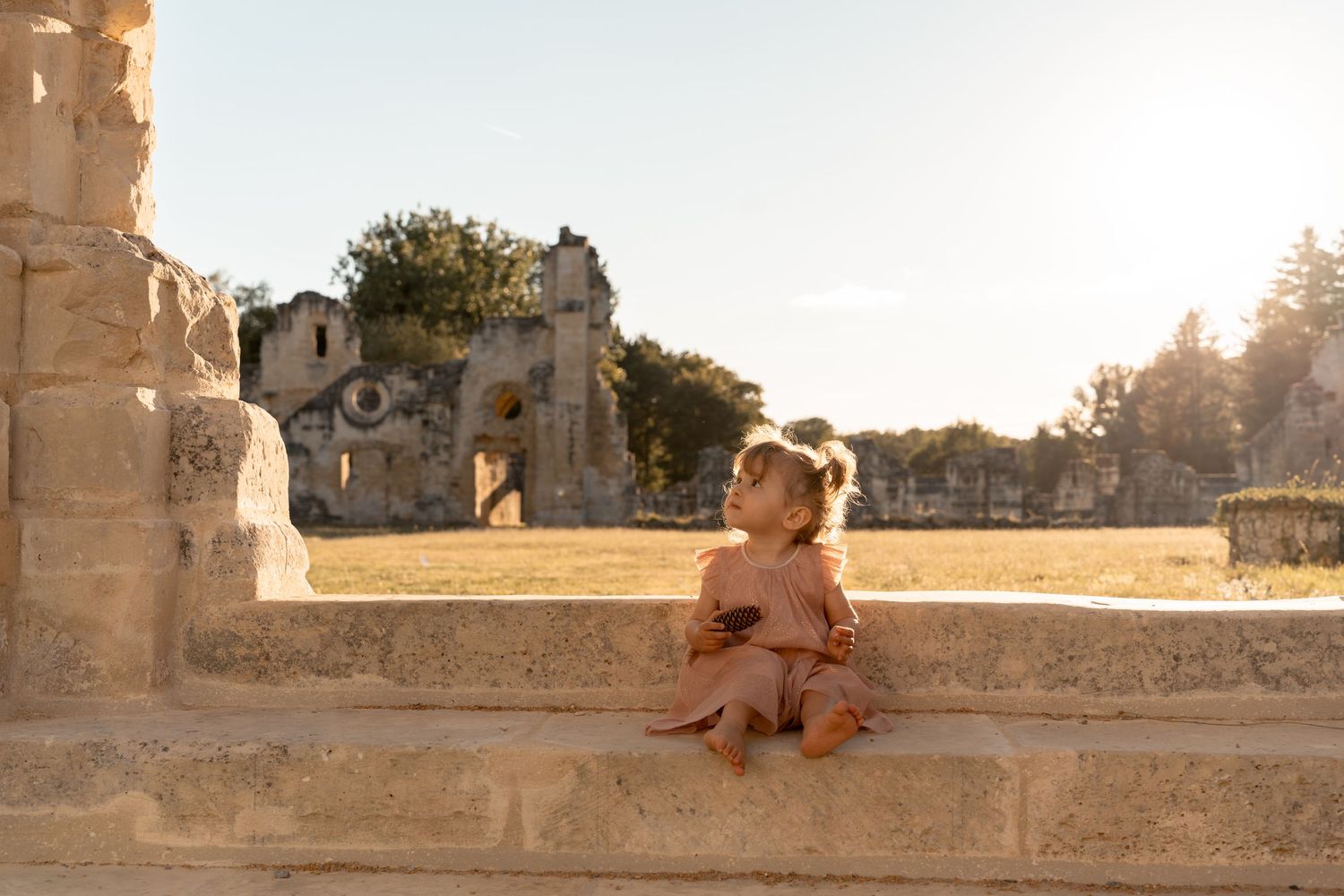 A toddler girl sits on ancient stone steps at sunset, gazing up at historic ruins in warm golden light.