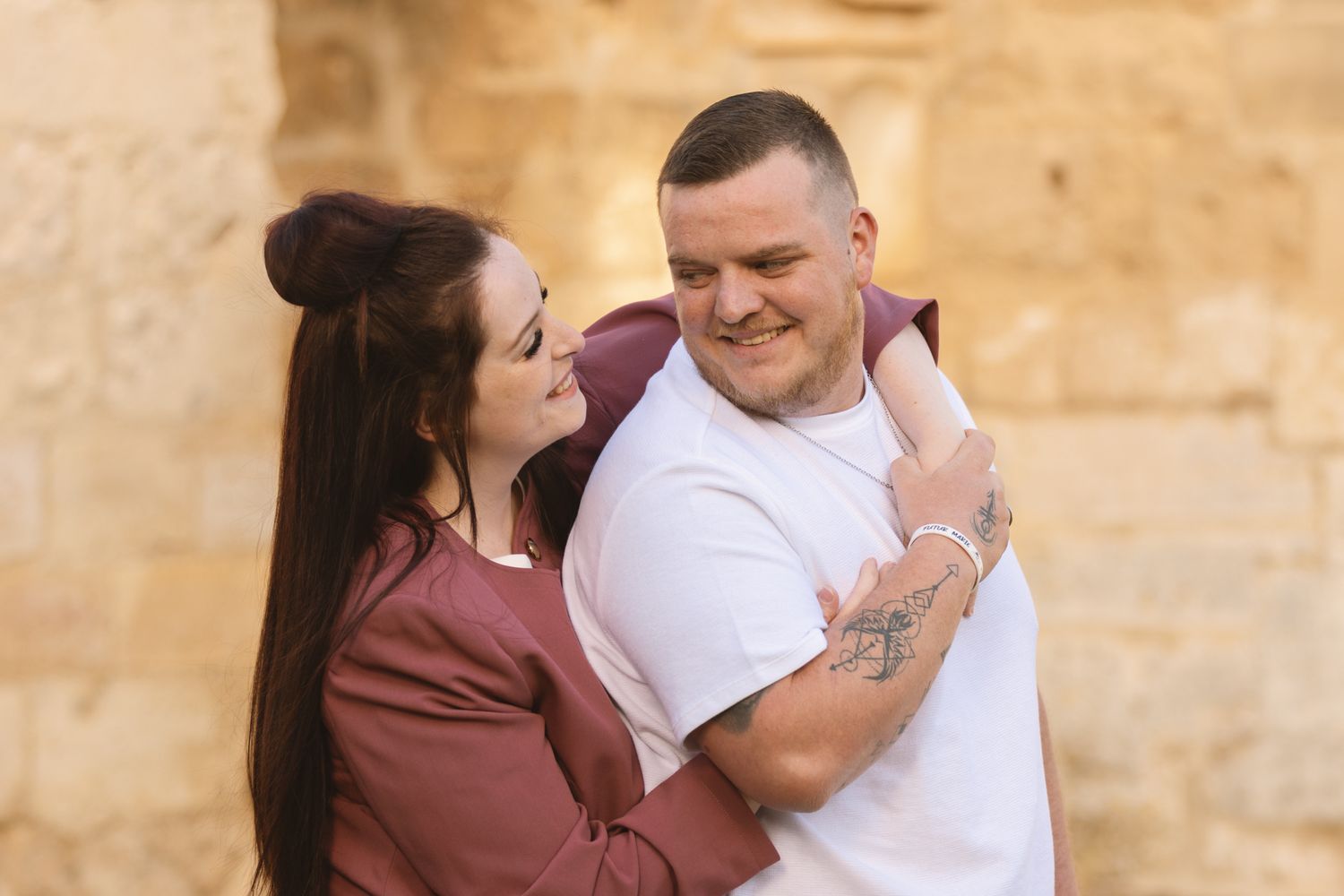 A couple embracing and smiling at each other outdoors, the woman in burgundy hugging a man in a white t-shirt.