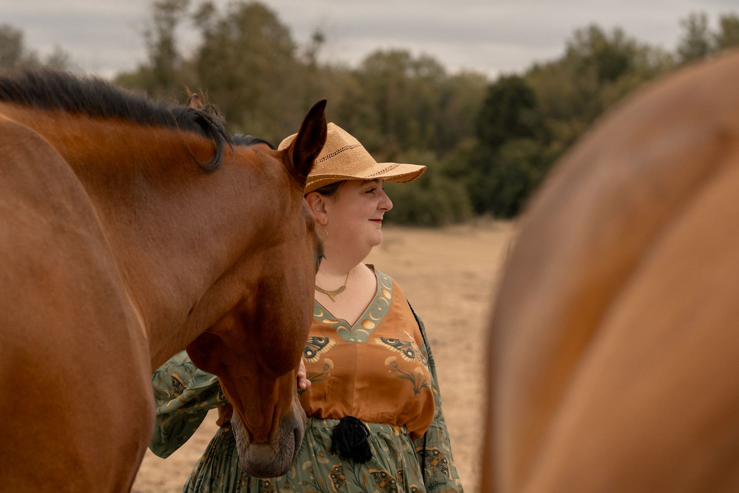 Woman in vintage attire with yellow hat stands between two brown horses in a sunny field.