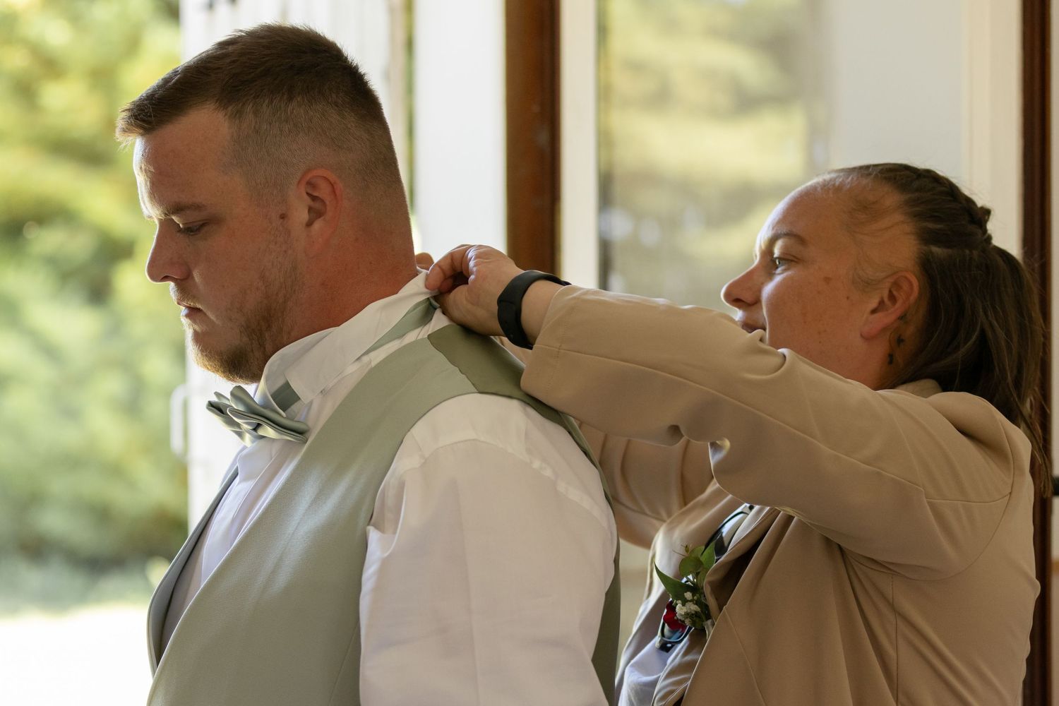 Woman helping groom adjust his sage green vest before wedding ceremony indoors.