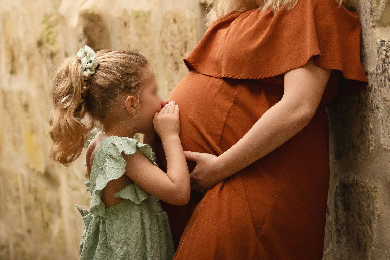 A young girl in a green dress kisses a pregnant woman's belly while holding hands outdoors.