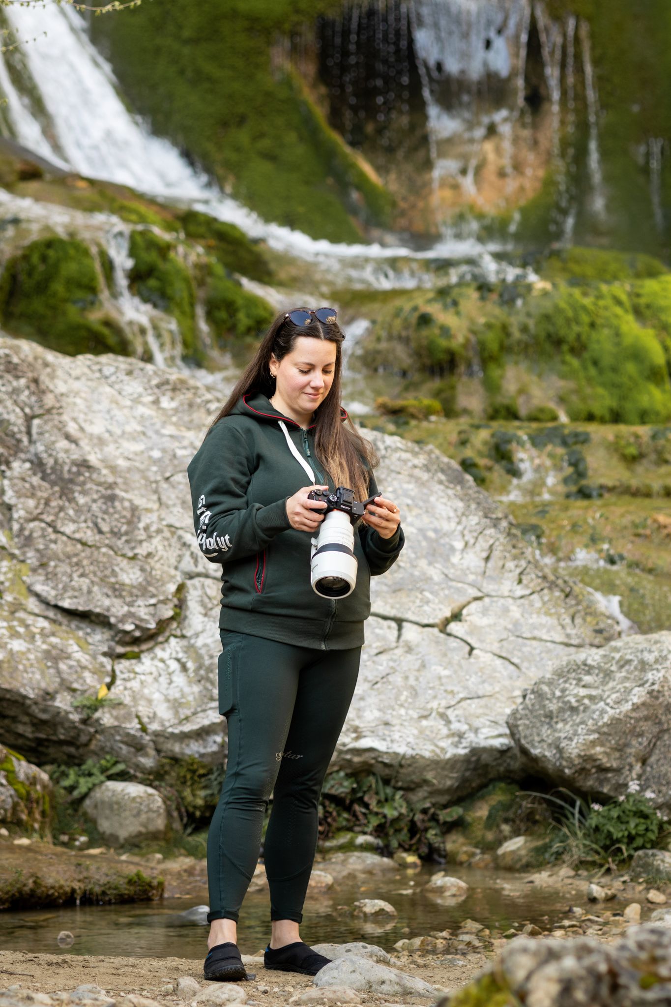A woman photographer reviews images on her camera near a scenic waterfall surrounded by mossy rocks.