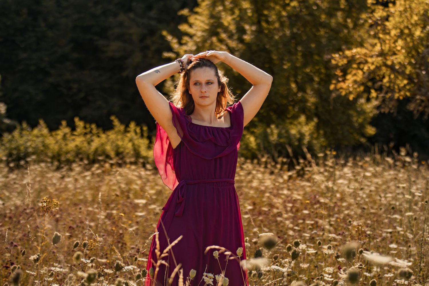 Woman in burgundy dress posing in golden field with trees in background during sunset.