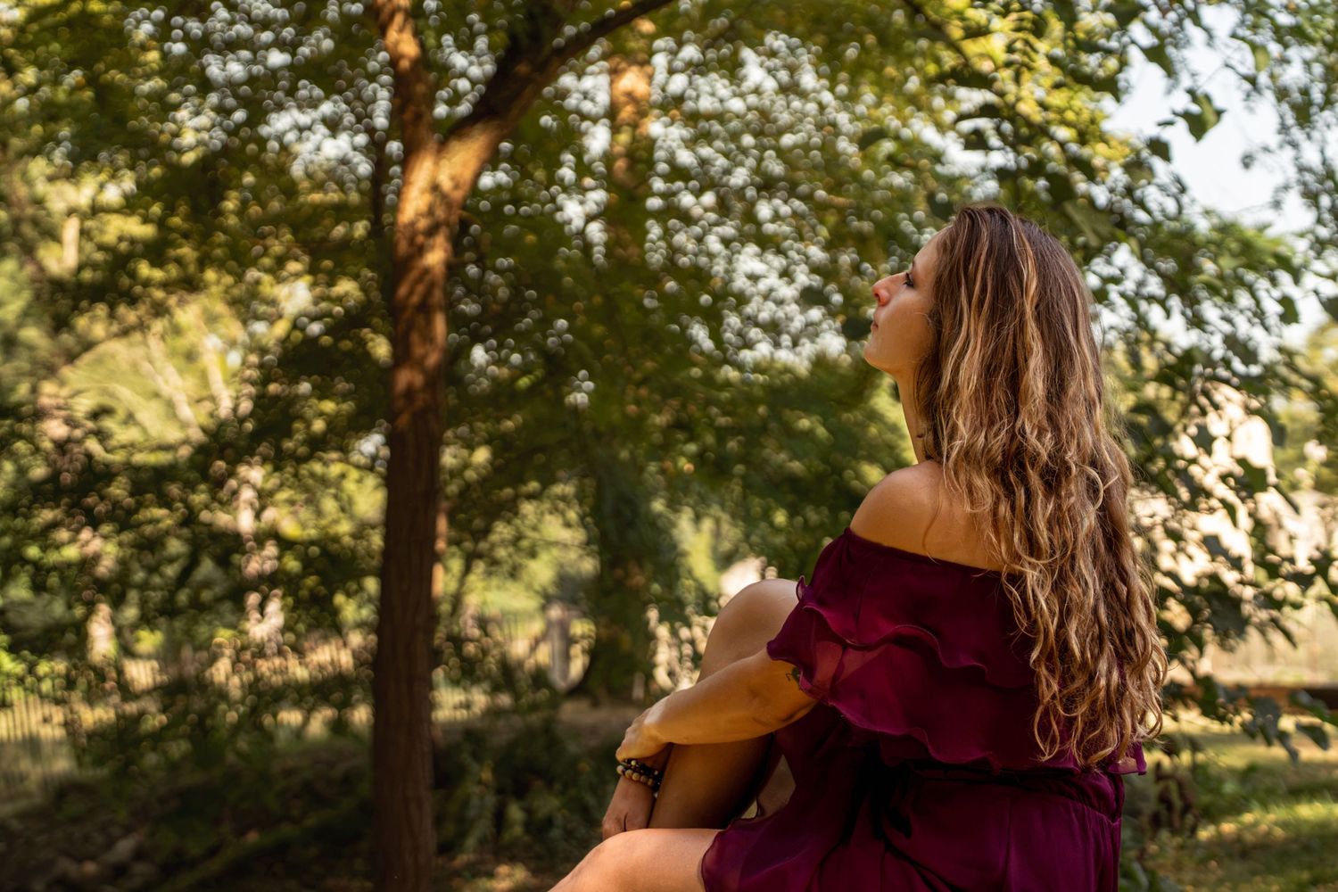 Woman in burgundy off-shoulder dress playing ukulele outdoors among lush green trees in warm sunlight.