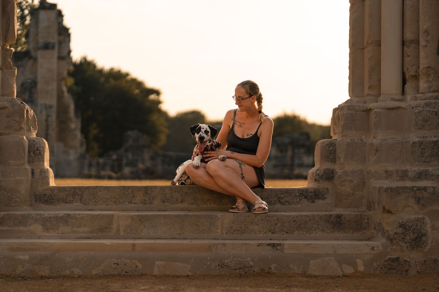 Woman sitting on ancient stone steps at sunset, holding a Dalmatian dog, surrounded by historic ruins.