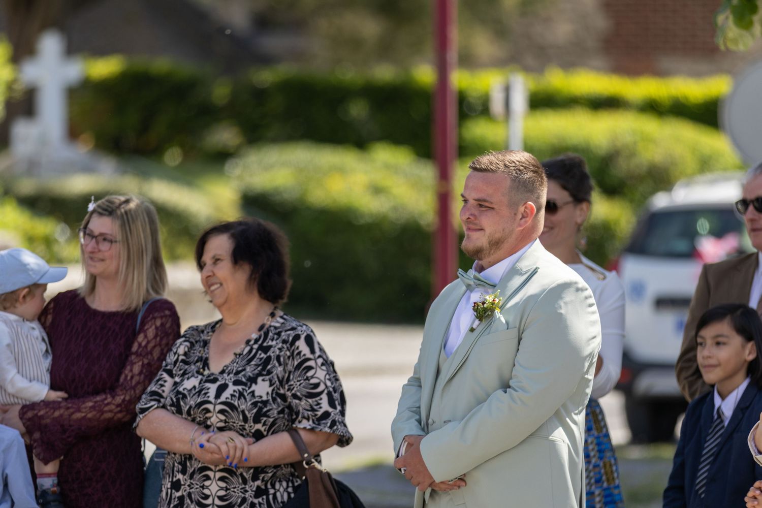 Groom in light suit with boutonniere stands at outdoor wedding ceremony surrounded by guests in sunny weather.