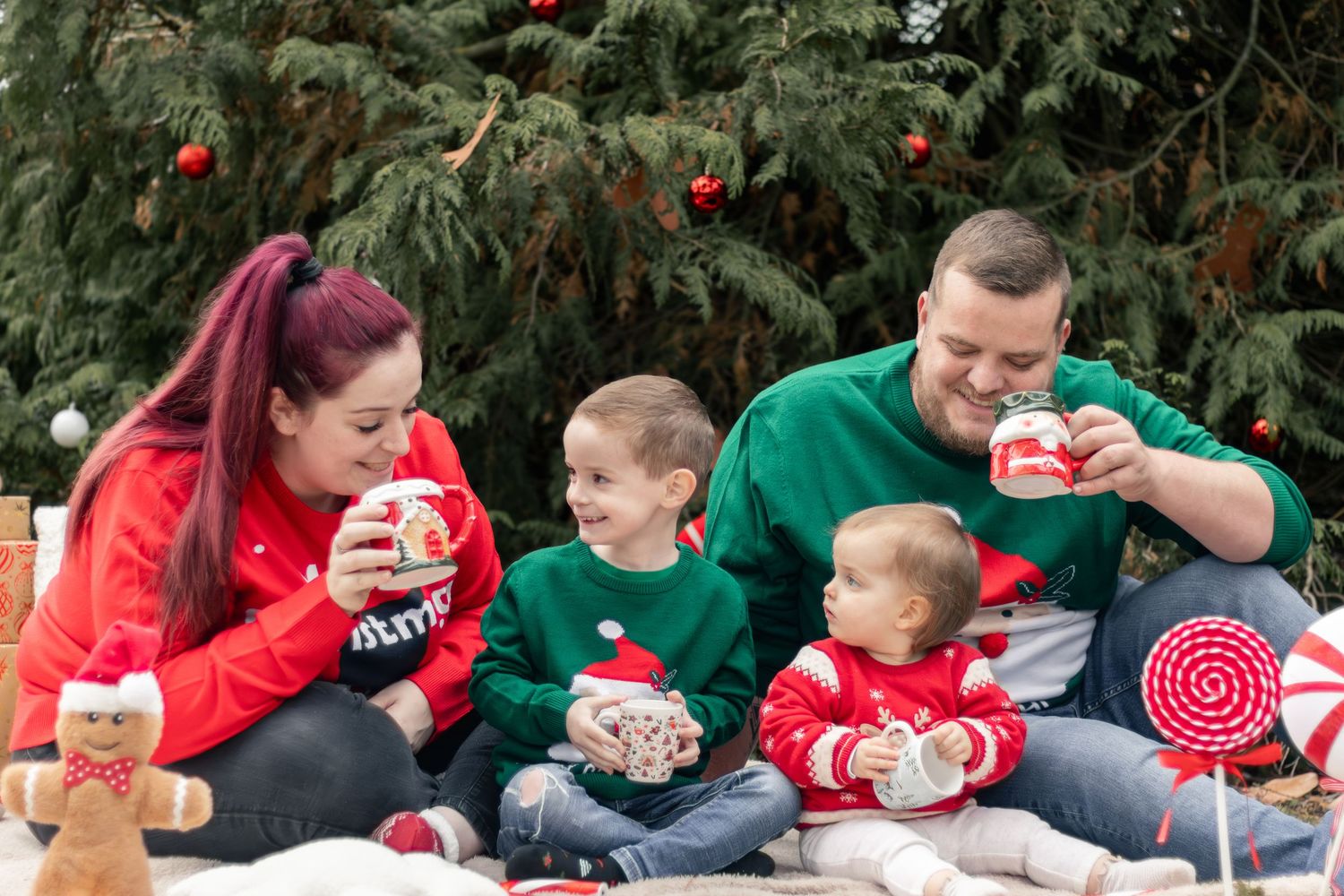 Family of four in Christmas sweaters enjoying hot cocoa and candy canes outdoors near a decorated tree.