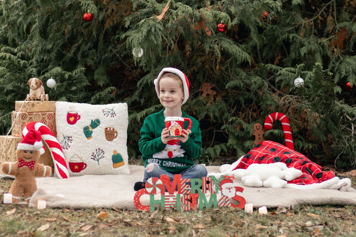 A young child in a Santa hat sits among festive Christmas decorations outdoors near a tree.