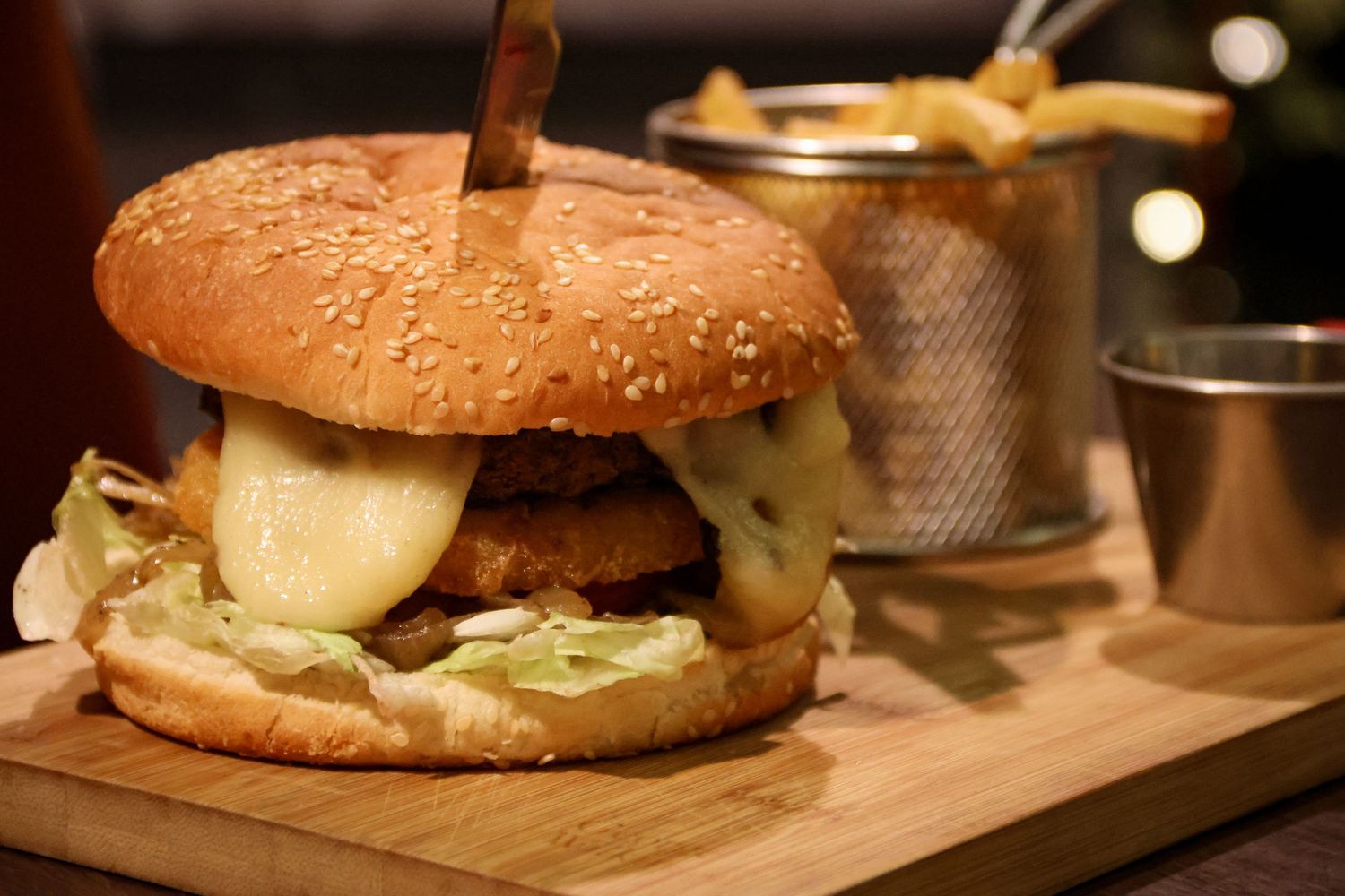 A gourmet cheeseburger with lettuce on a wooden board served with crispy french fries in a metal basket.