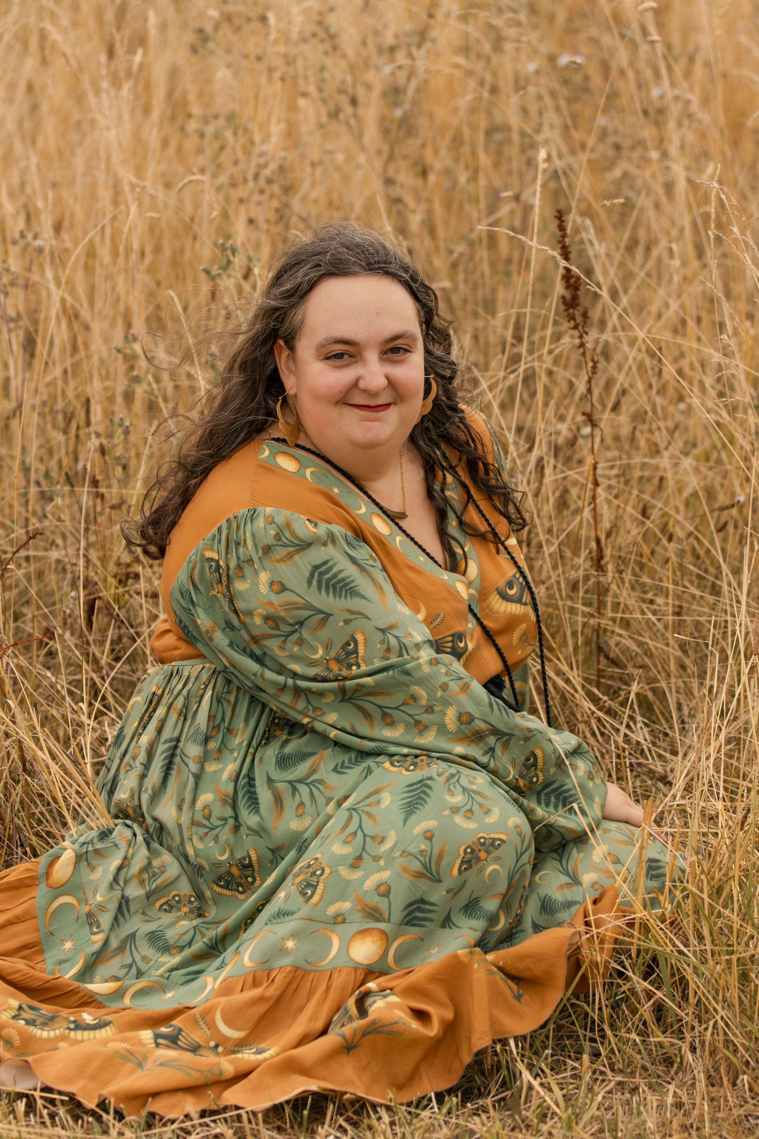 Woman in green and gold floral dress sitting in golden wheat field, smiling at camera.