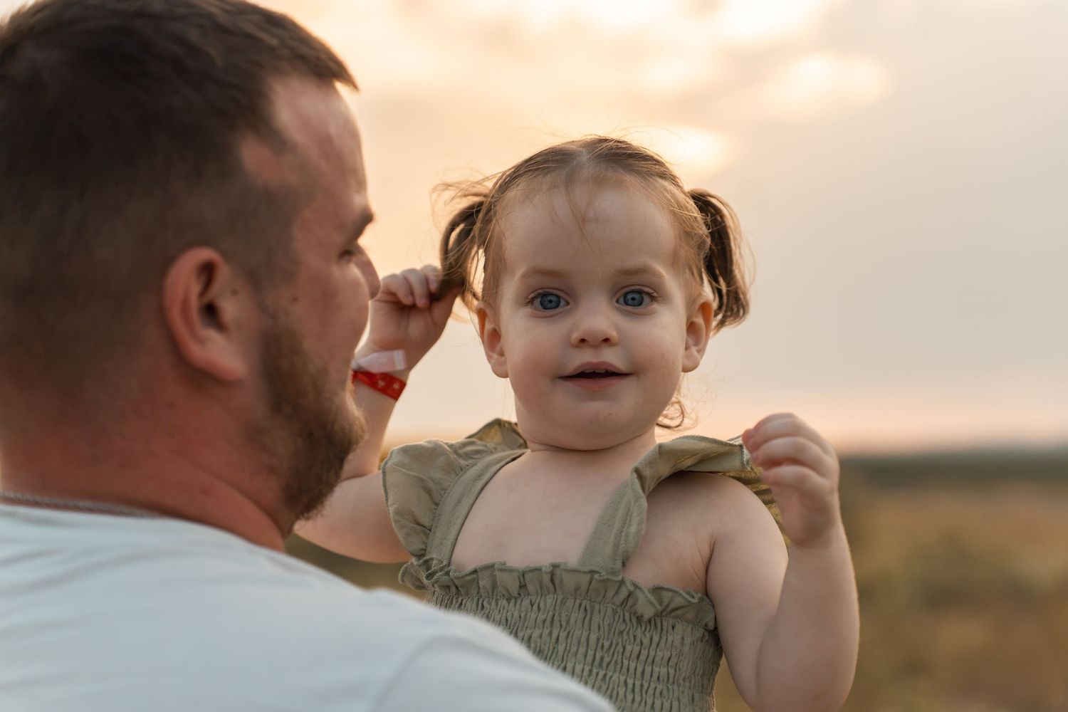 Father holding smiling baby girl with pigtails outdoors at sunset in a warm family moment.