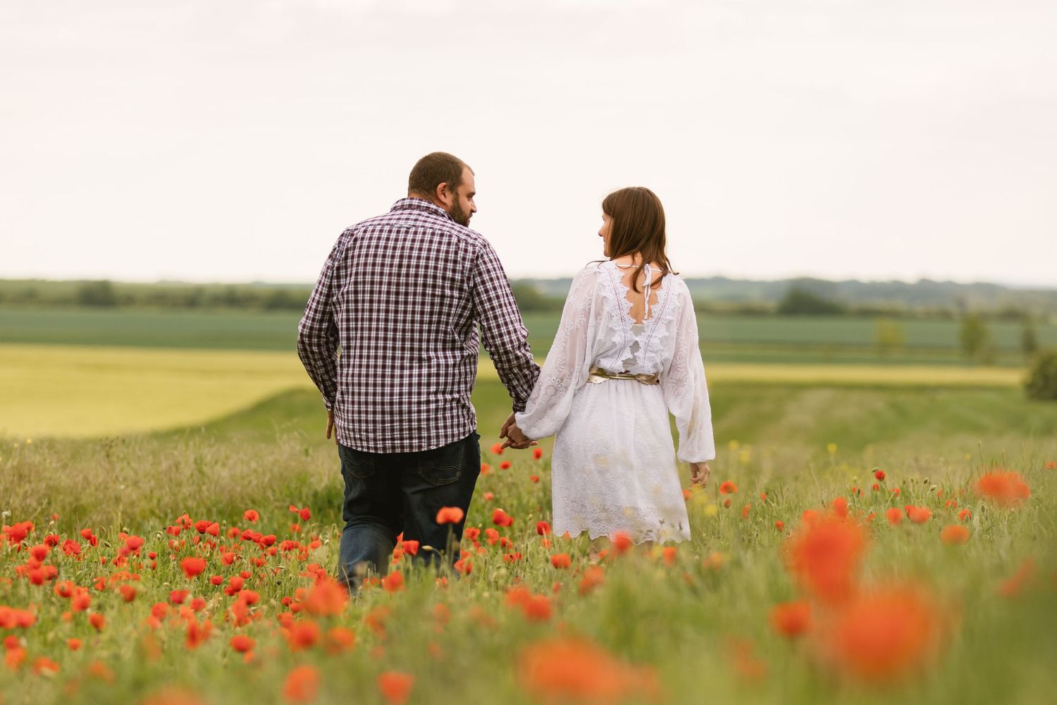 Couple holding hands walking through a blooming red poppy field on a cloudy day.