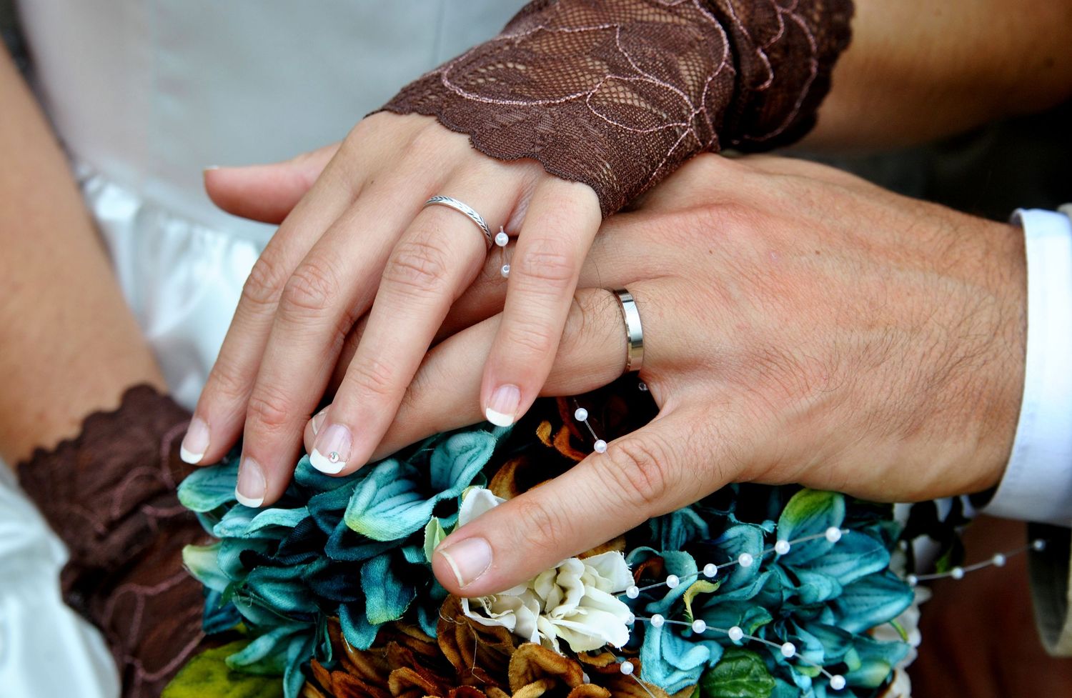 Newlyweds displaying wedding rings on hands resting over a teal and brown floral bouquet.