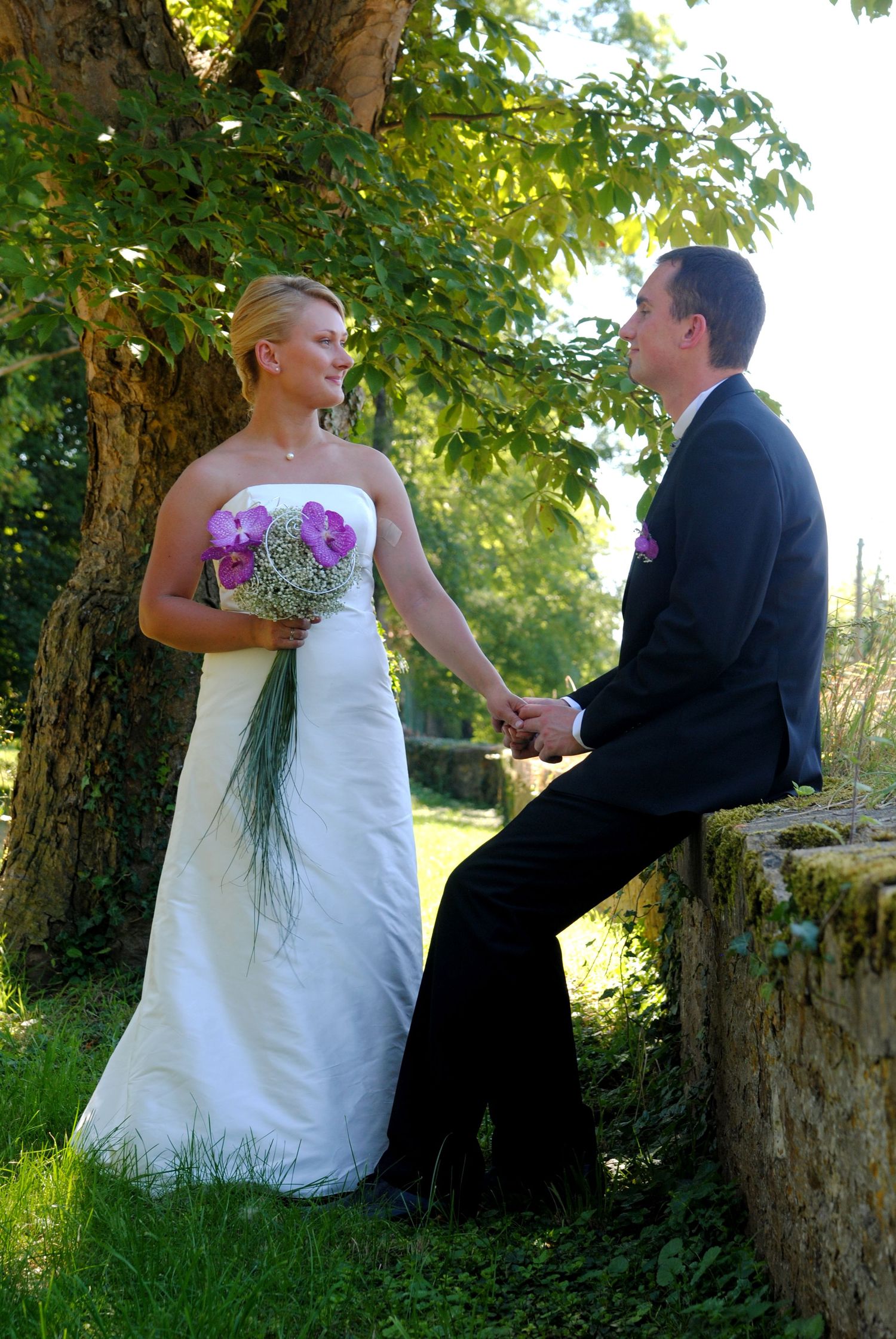 Bride in white strapless gown holding purple bouquet holds hands with groom in black suit outdoors near tree.