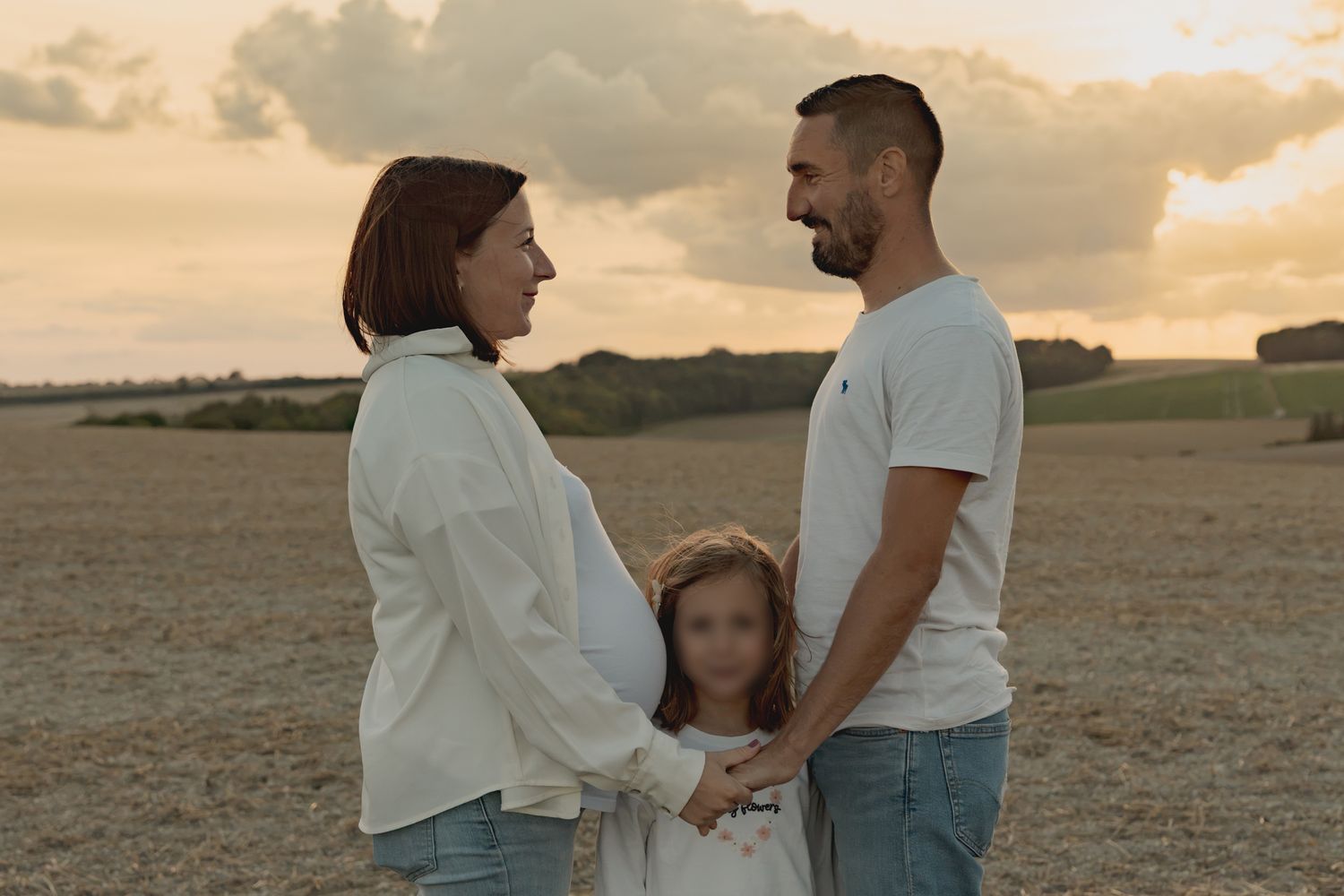 Pregnant couple holding hands with young daughter in golden field at sunset.