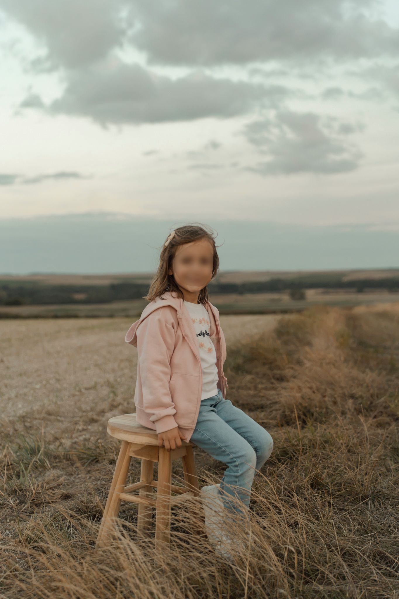 Young girl in pink hoodie and jeans sitting on wooden stool in golden field under cloudy sky.