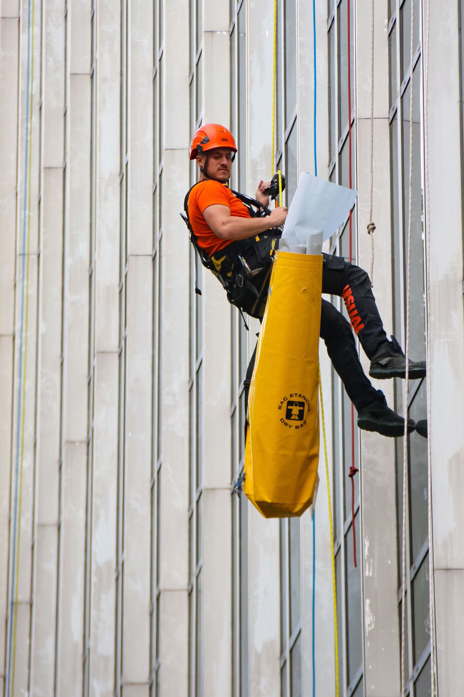 Industrial rope access worker in orange helmet and harness descending a building facade with yellow equipment bag.