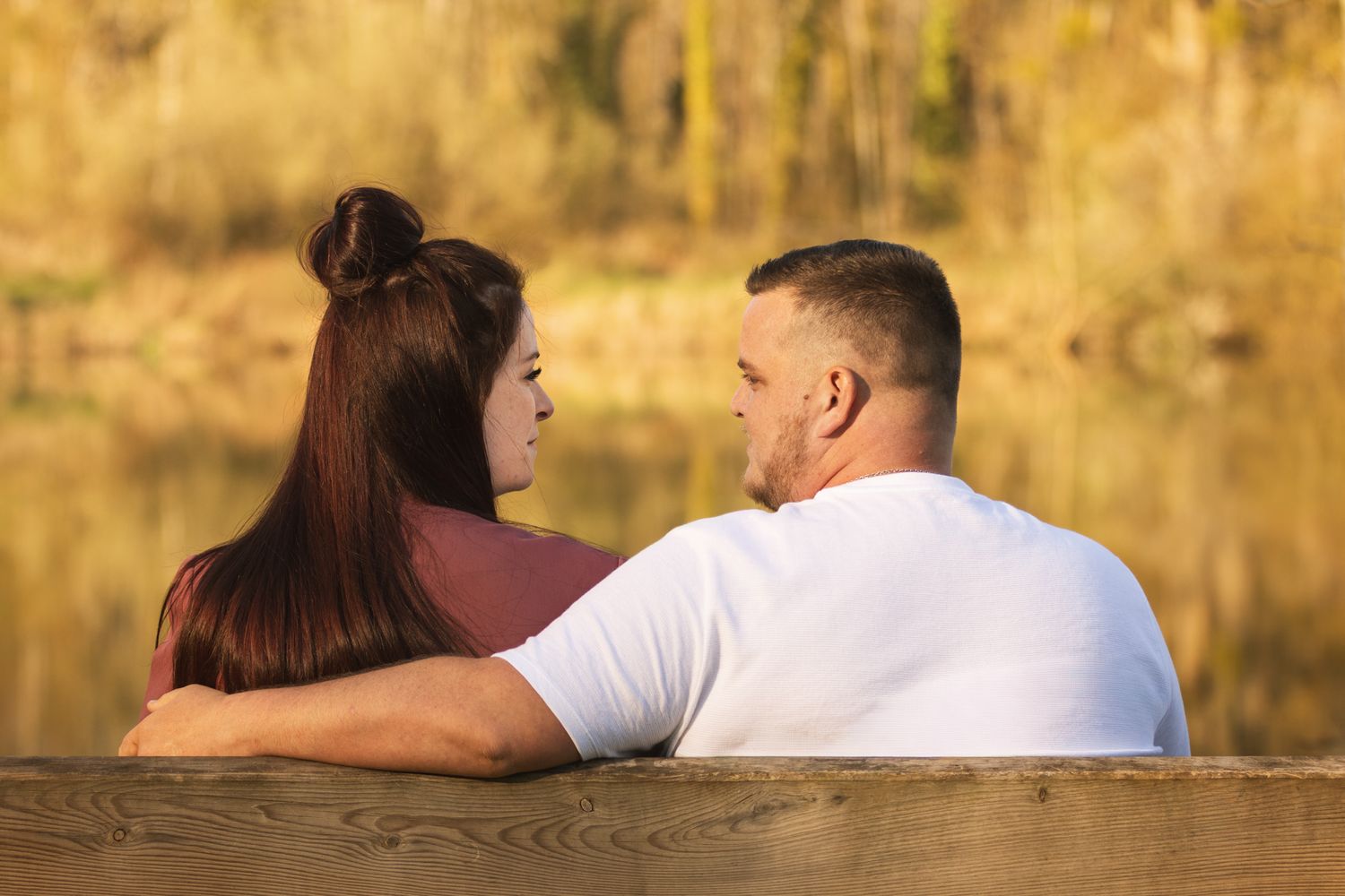 Couple sitting on a wooden dock by a lake, facing each other with autumn foliage in the background.