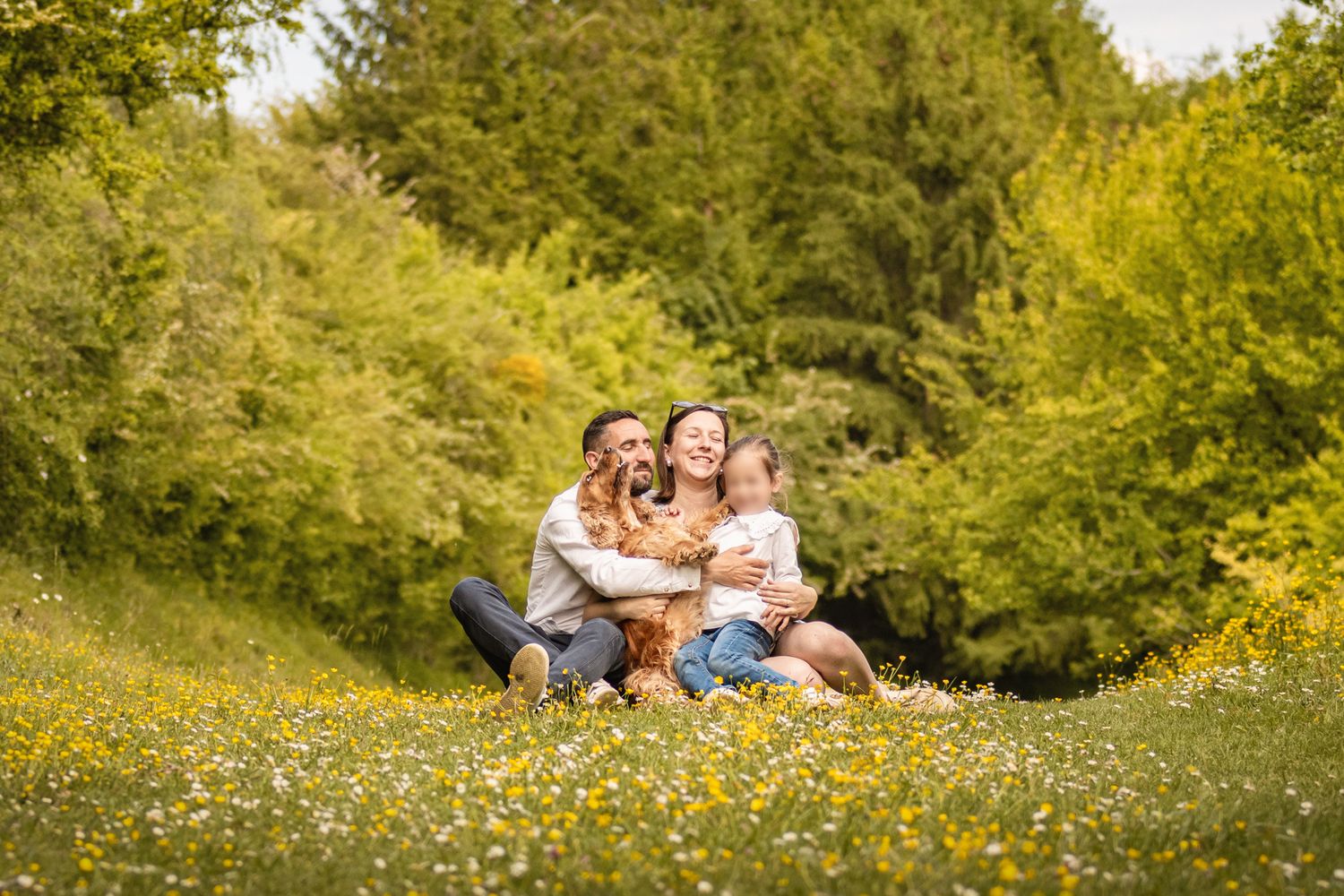 Happy family with dog sitting in a sunny wildflower meadow surrounded by lush green trees.