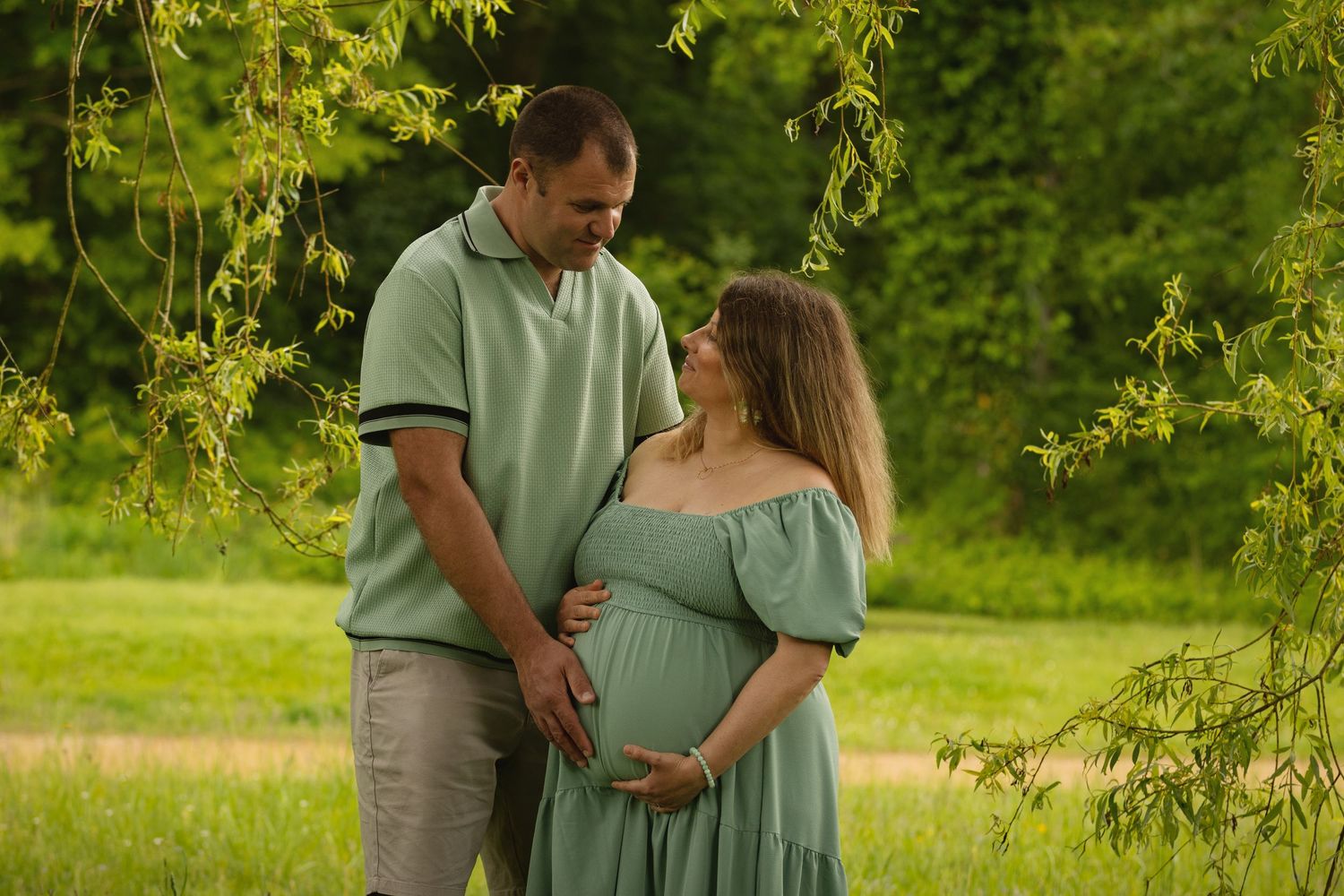 Couple in matching green outfits sharing a tender moment outdoors, with man gently holding pregnant woman's belly.