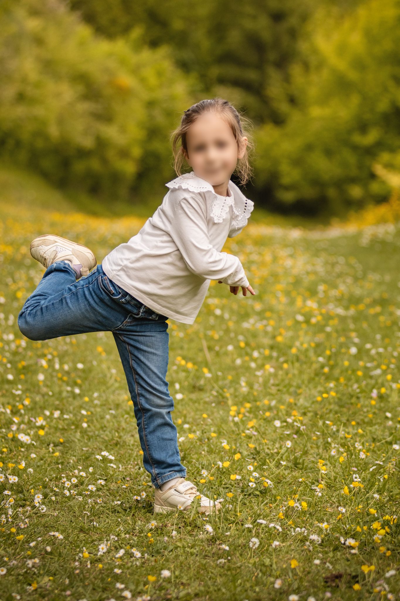 Young girl balancing on one leg in a flower-filled meadow, wearing jeans and white top on a sunny day.