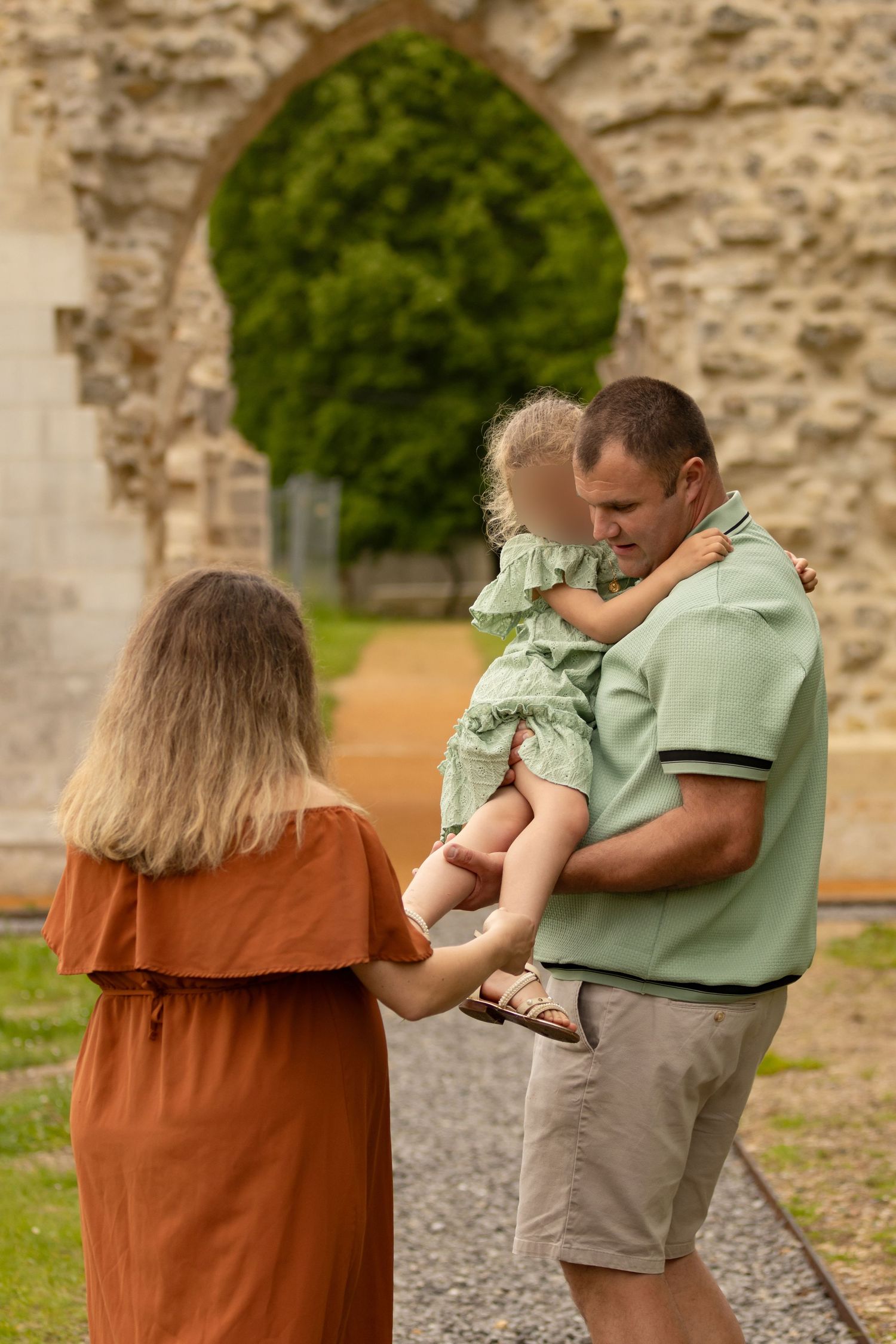 A family of three stands before a stone arch, father holding toddler in green outfit, mother in rust dress.