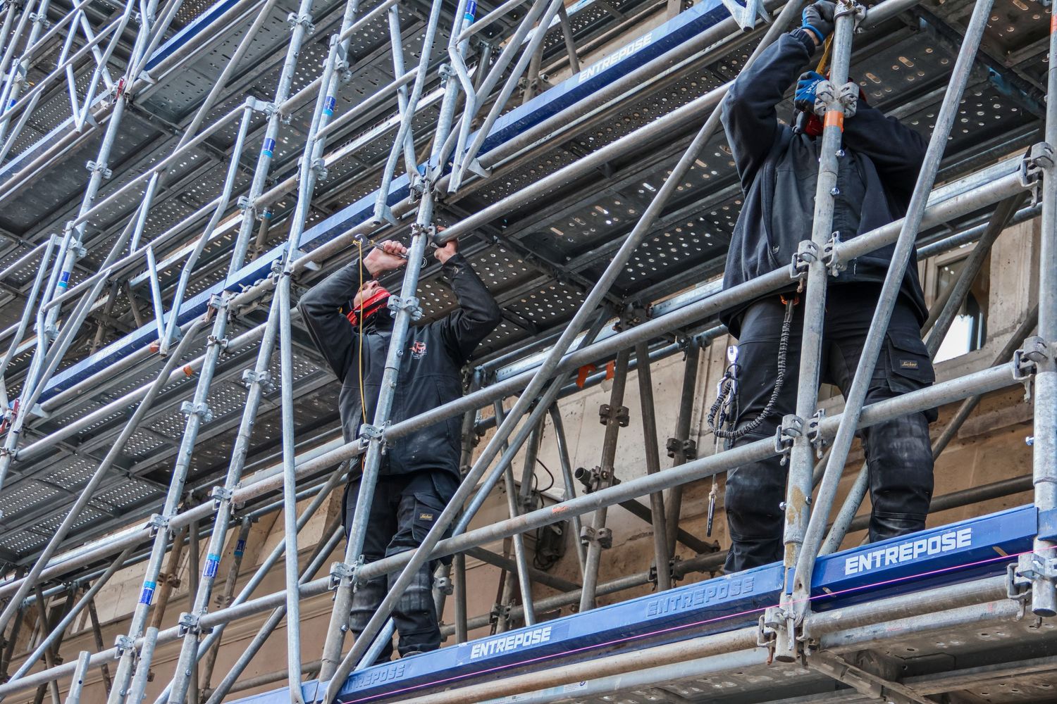 Two construction workers assembling metal scaffolding on a building facade during renovation work.