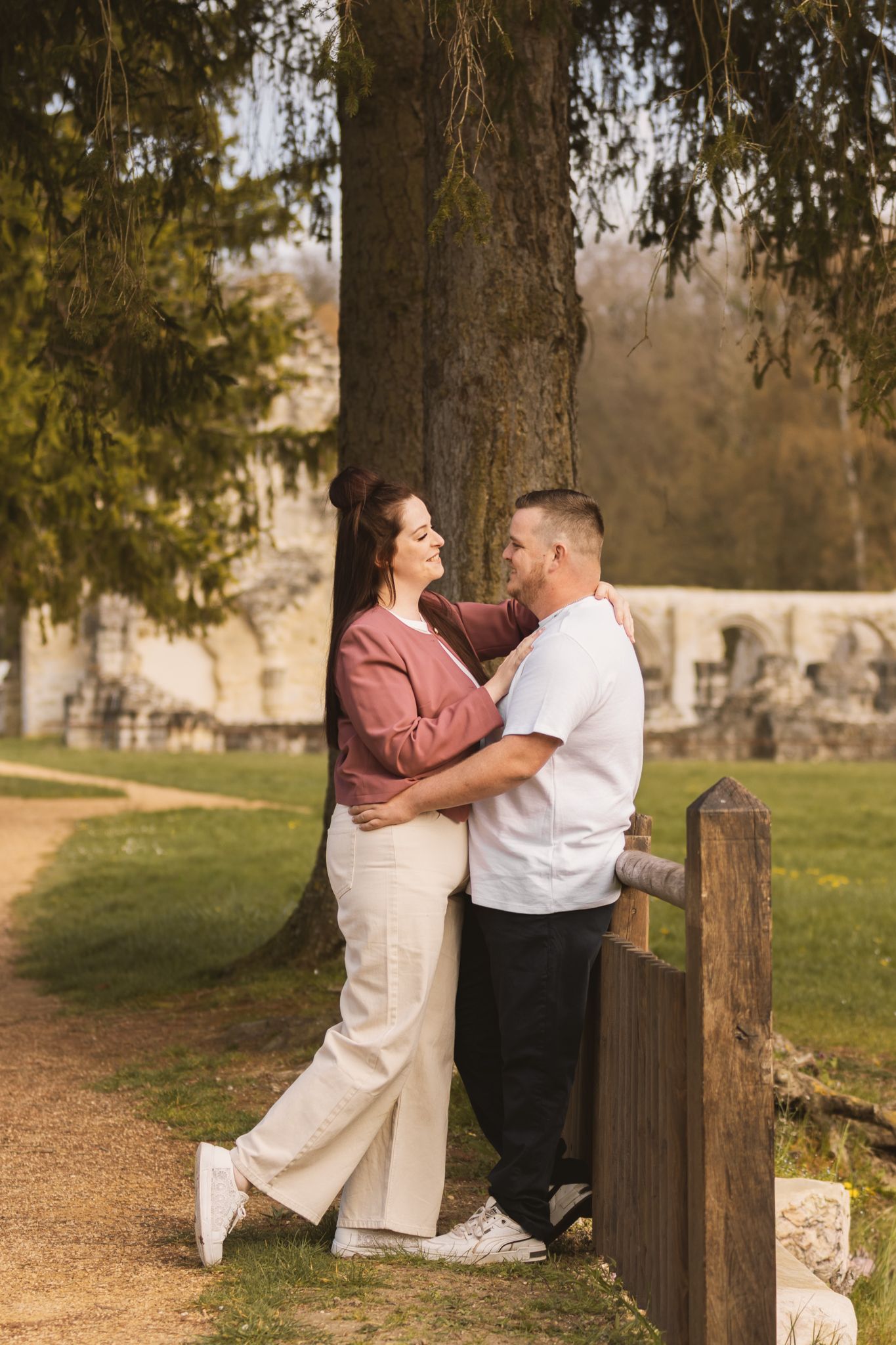 Couple embracing romantically by a large tree in a sunny park, woman in pink jacket and white pants, man in white shirt.