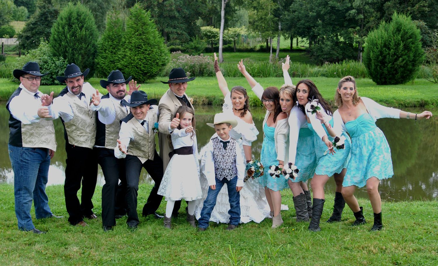 Western wedding party celebrating outdoors near a pond, groomsmen in cowboy hats and bridesmaids in teal dresses.
