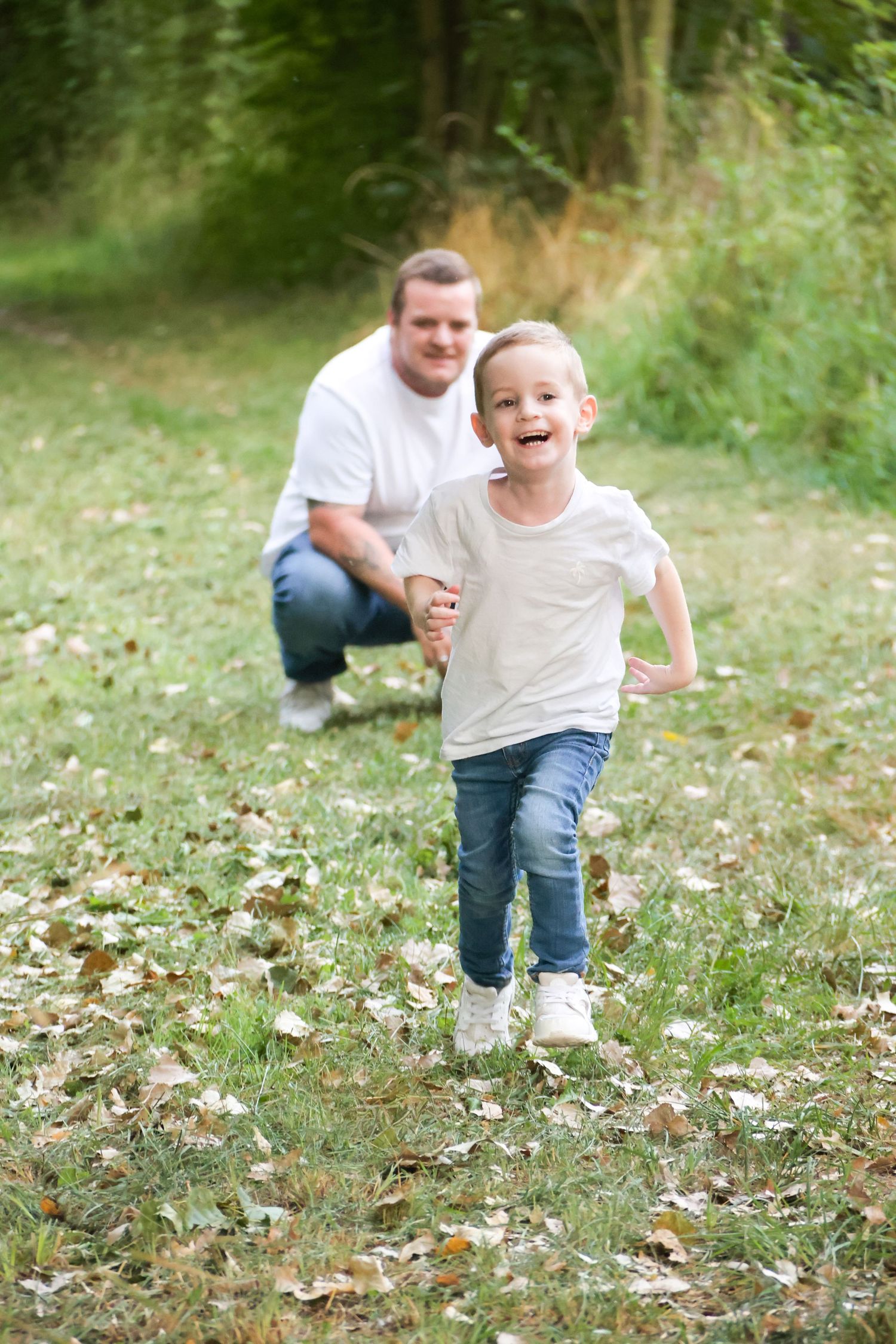 Happy young boy in white t-shirt and jeans running toward camera while father crouches in background in park.
