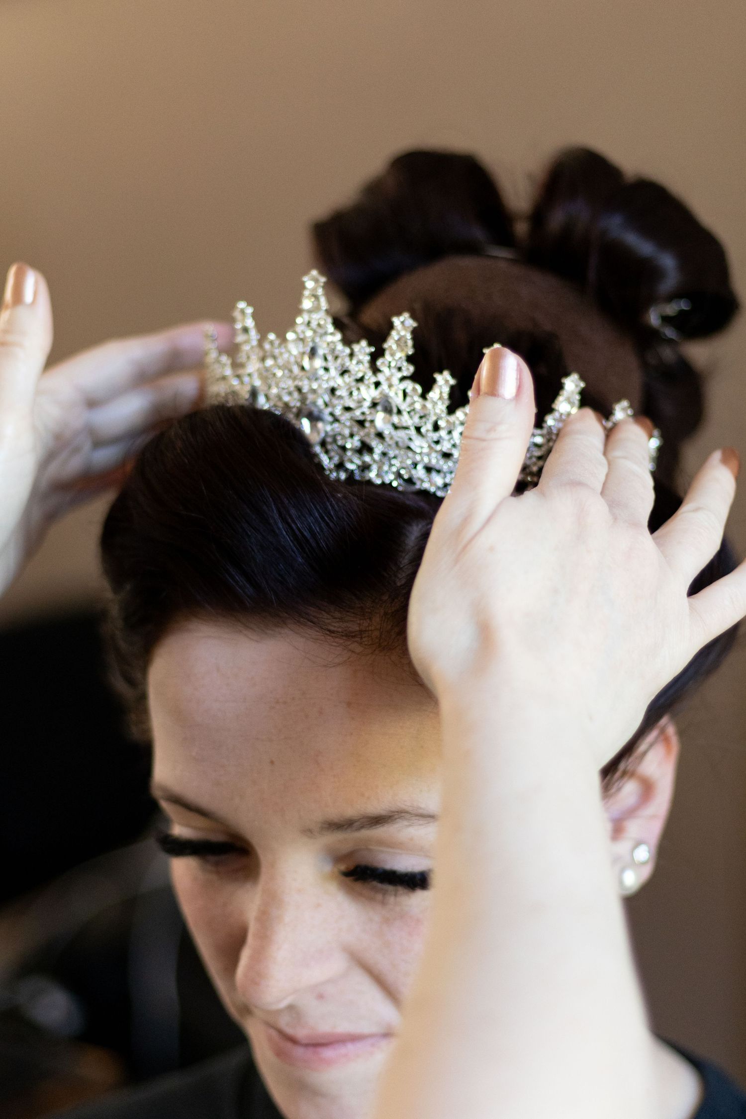 A bride having a sparkling tiara placed on her dark updo hairstyle by a stylist during wedding preparations.
