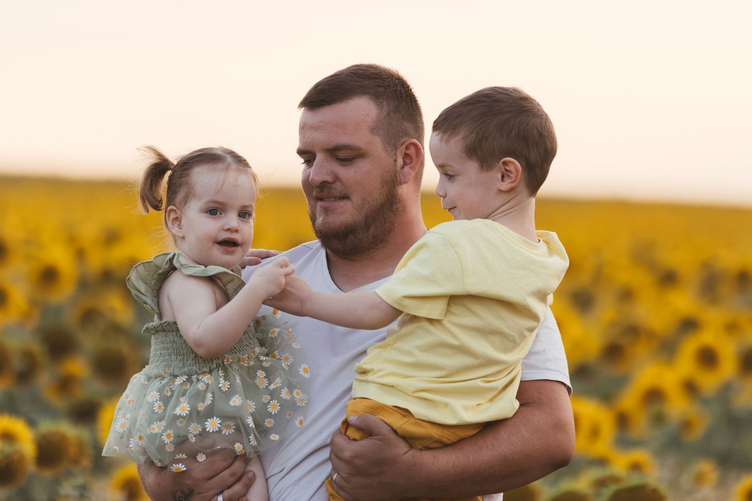 Father holding two young children in a sunflower field at sunset, smiling outdoors.