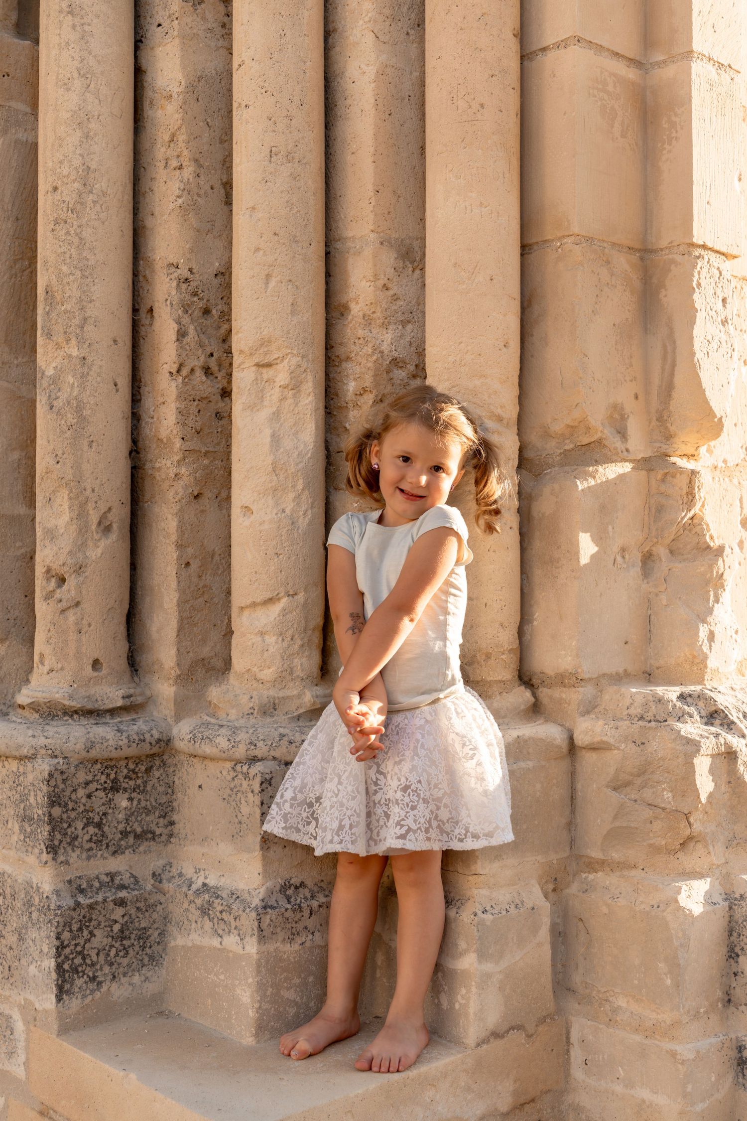 A smiling barefoot little girl in a white lace skirt leans against ancient stone columns.