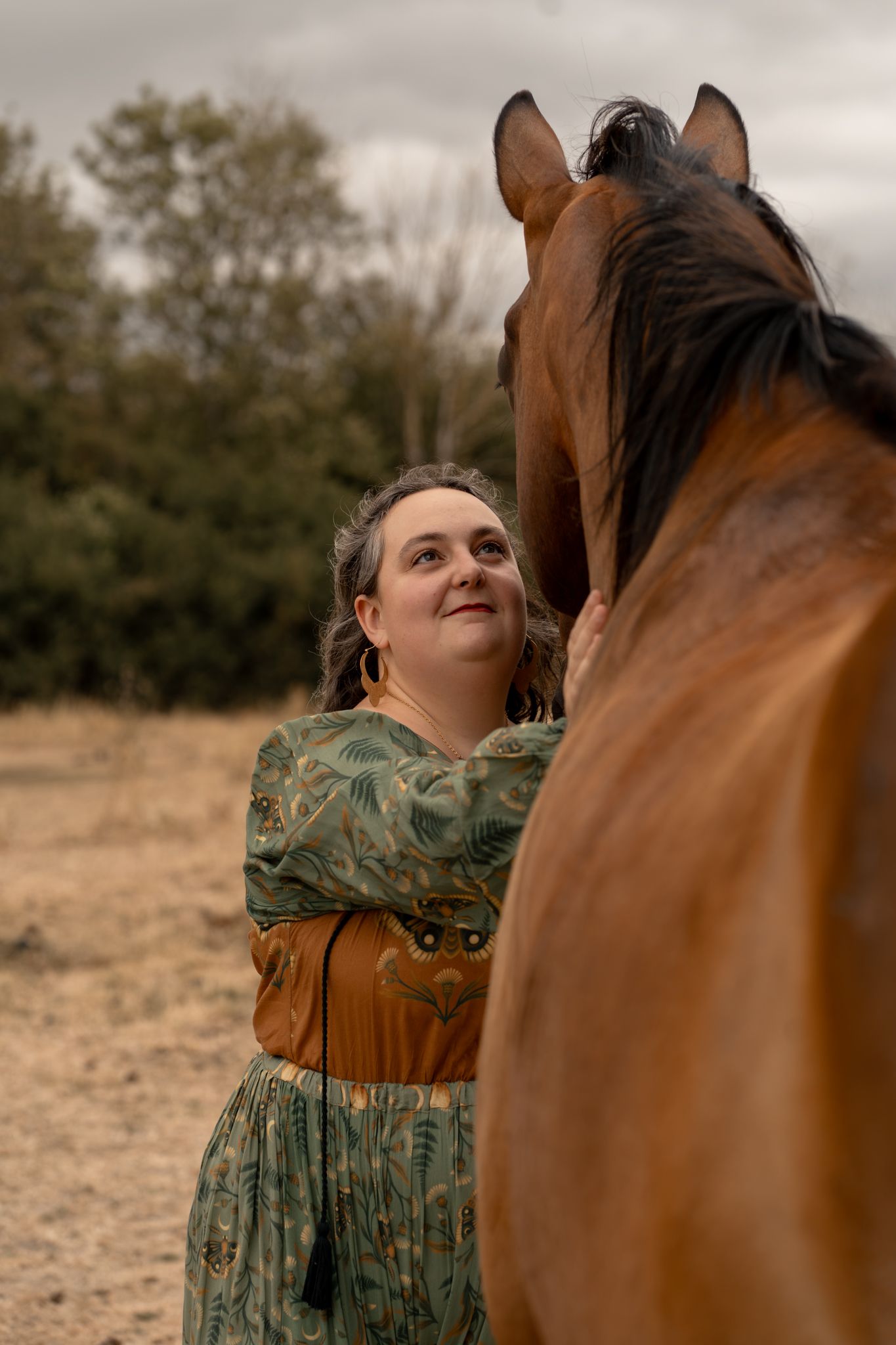 Woman in floral dress tenderly embracing a brown horse outdoors on a cloudy day.