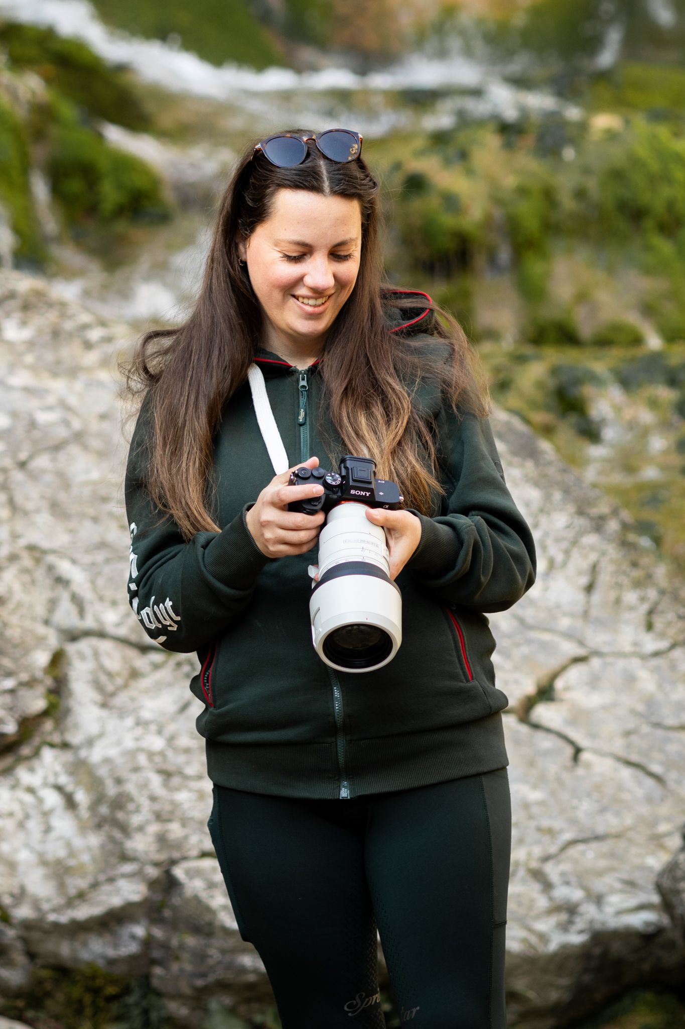 Smiling woman photographer holding a DSLR camera with telephoto lens outdoors near rocky terrain.