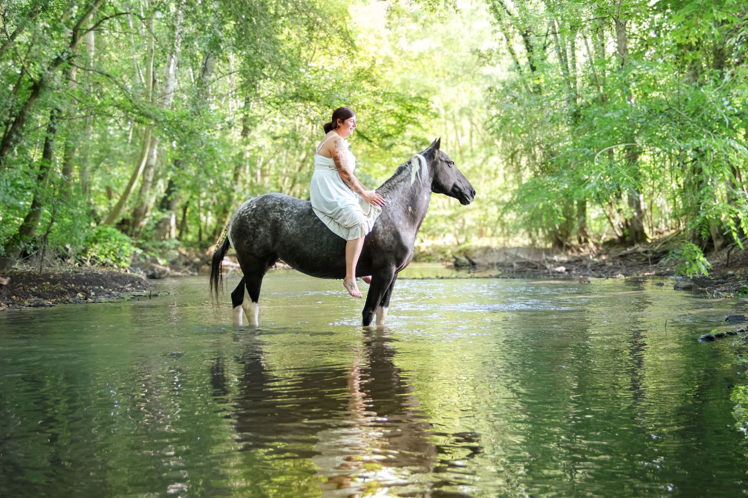 A woman in a white dress rides a grey horse through a serene shallow forest stream.